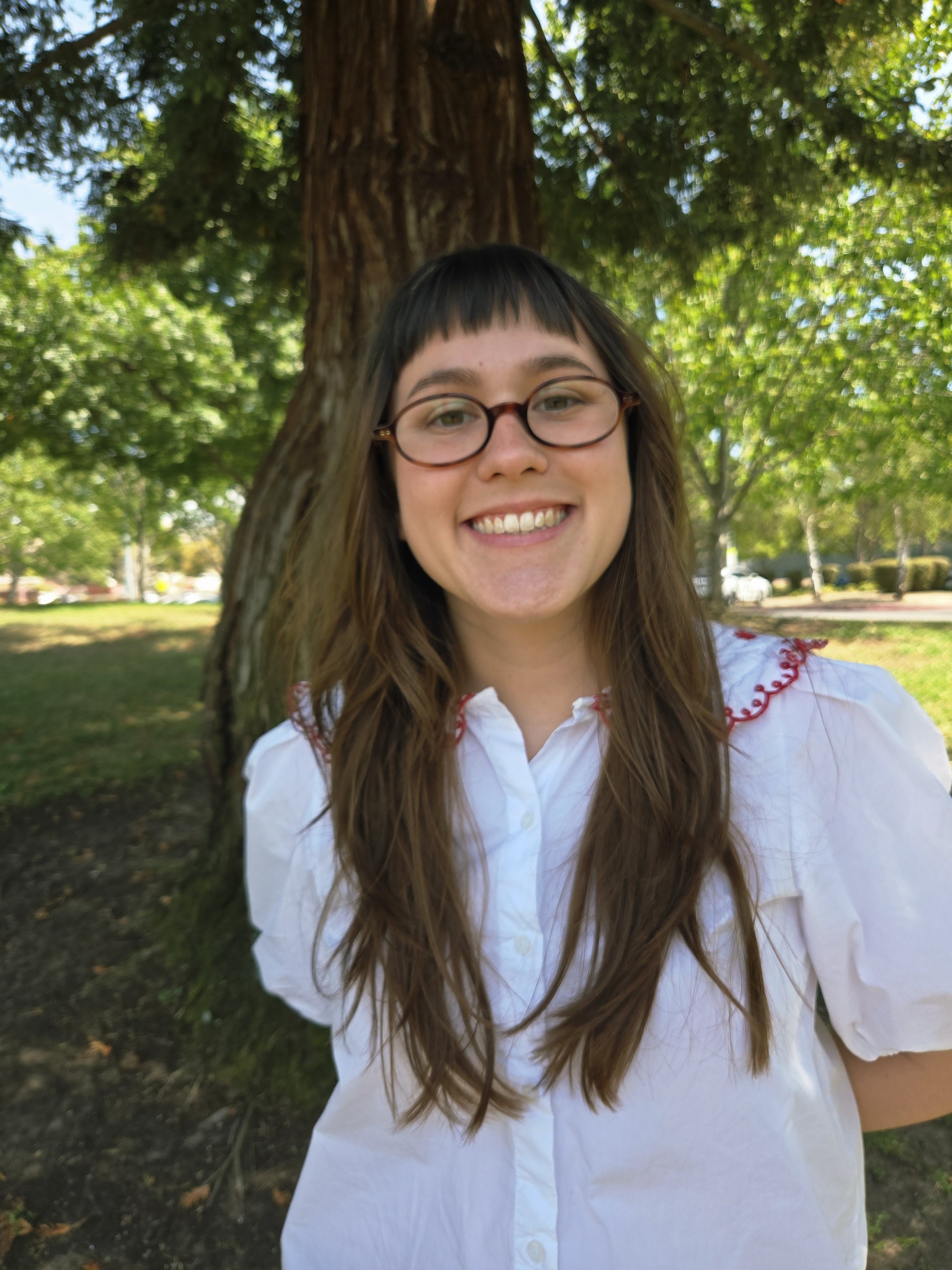 A young woman with long brown hair, glasses, and a white blouse smiling outside in front of a tree with green leaves.