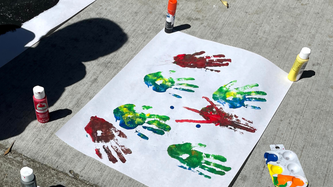 Colorful handprints on a white sheet of paper with paint bottles surrounding it on a concrete sidewalk.