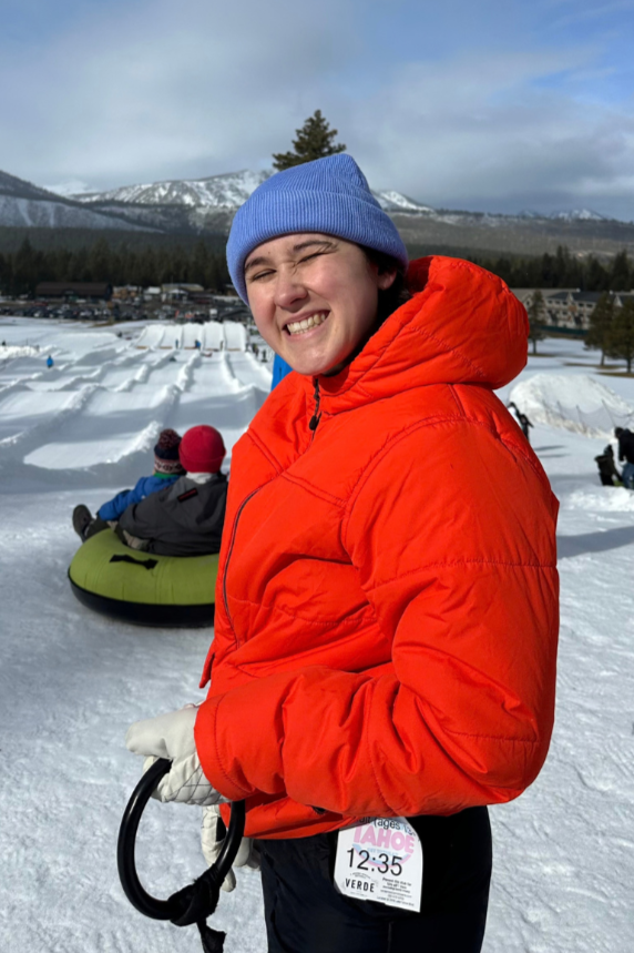 A smiling young person in an orange jacket and blue beanie standing in a snowy landscape, holding a leash with dogs in the background, with mountains and a cloudy sky.