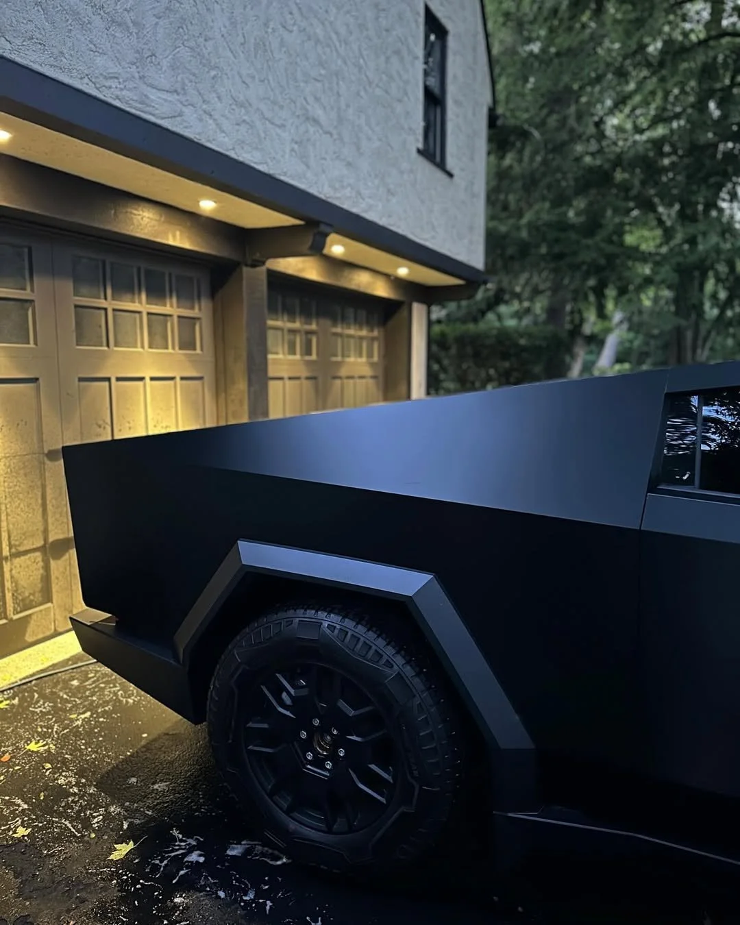 Close-up of the rear side of a matte black vehicle, parked in front of a garage with a modern design, under exterior lighting, with trees in the background.