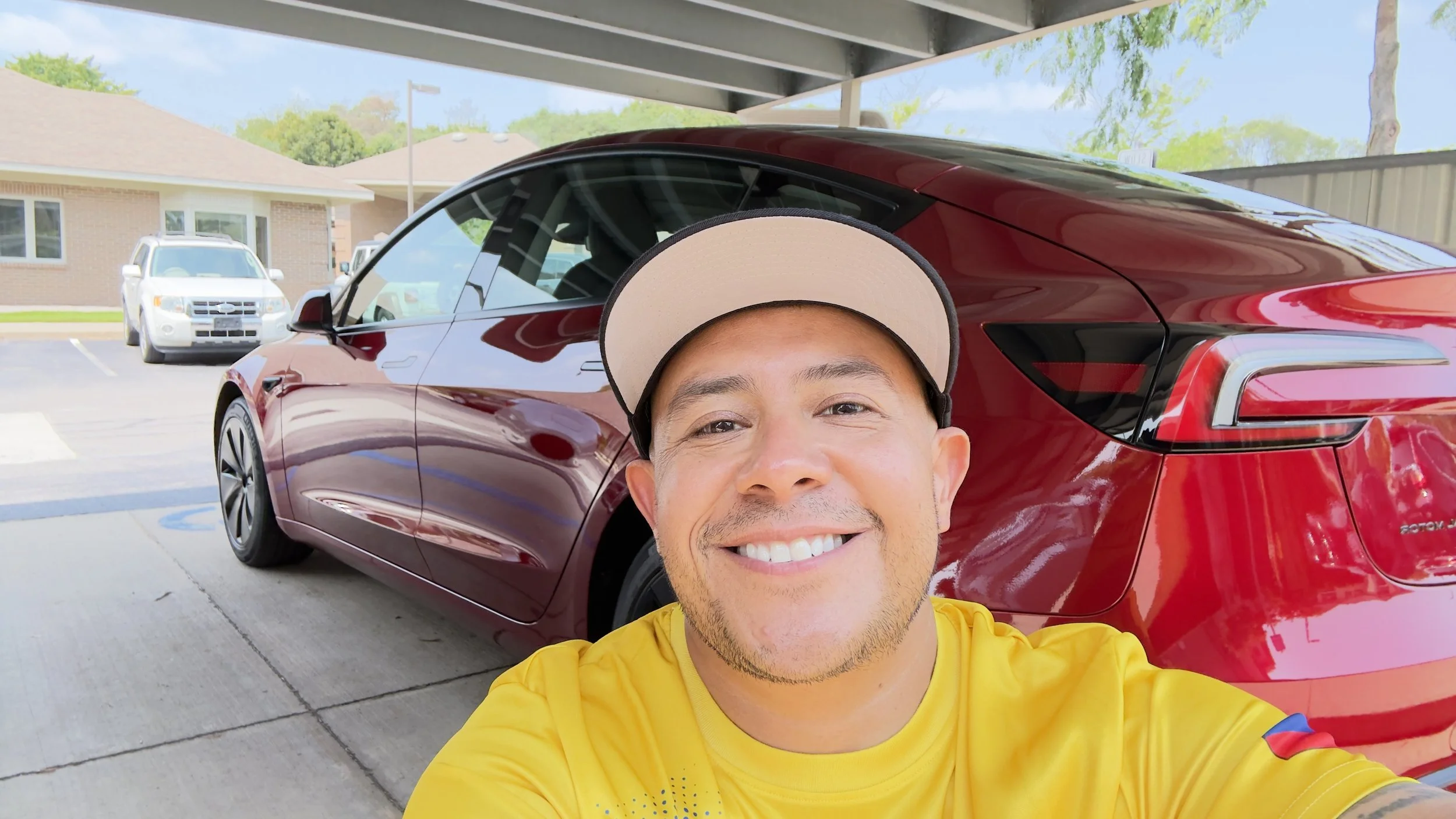 A man smiling in a yellow shirt taking a selfie in front of a red car in a parking lot, with other cars and buildings in the background.