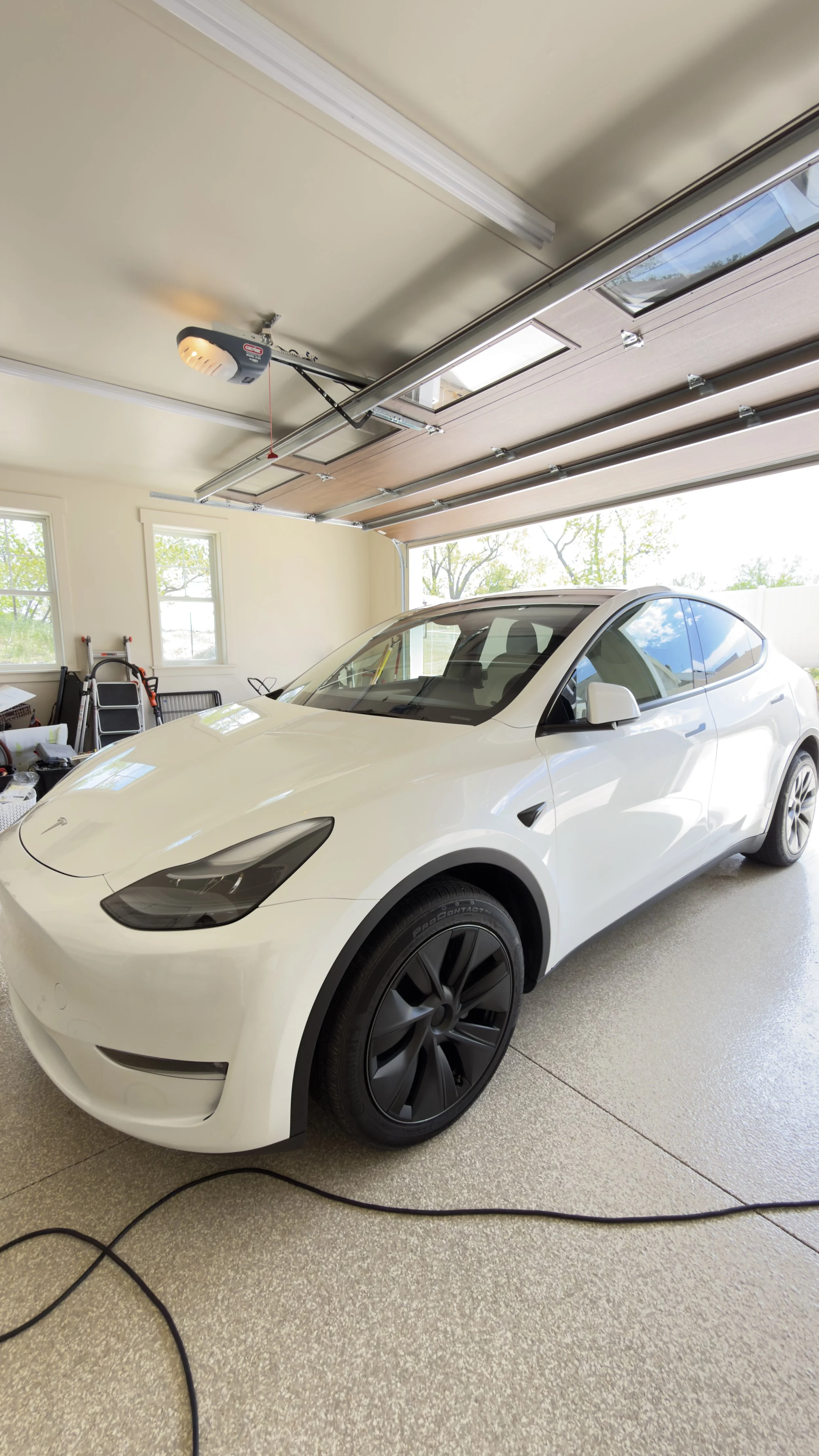 A white Tesla Model Y parked in a garage with the garage door partially open, revealing trees outside.