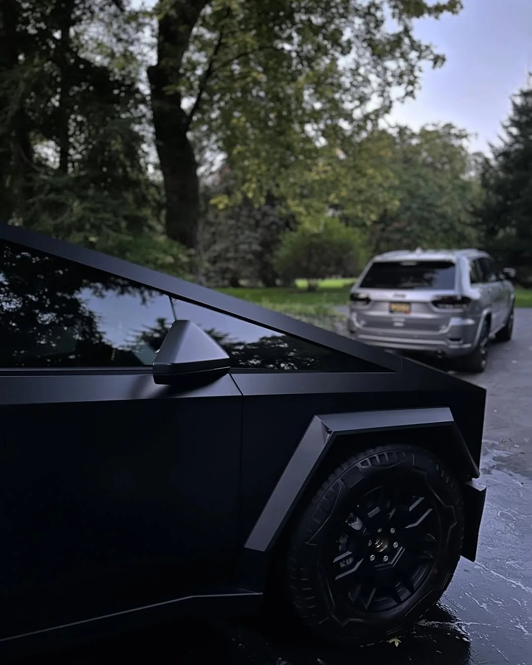 Close-up of the front left corner of a black modern car with a side mirror, parked outside on a wet driveway next to a gray SUV, in a lush green area with trees and a cloudy sky.