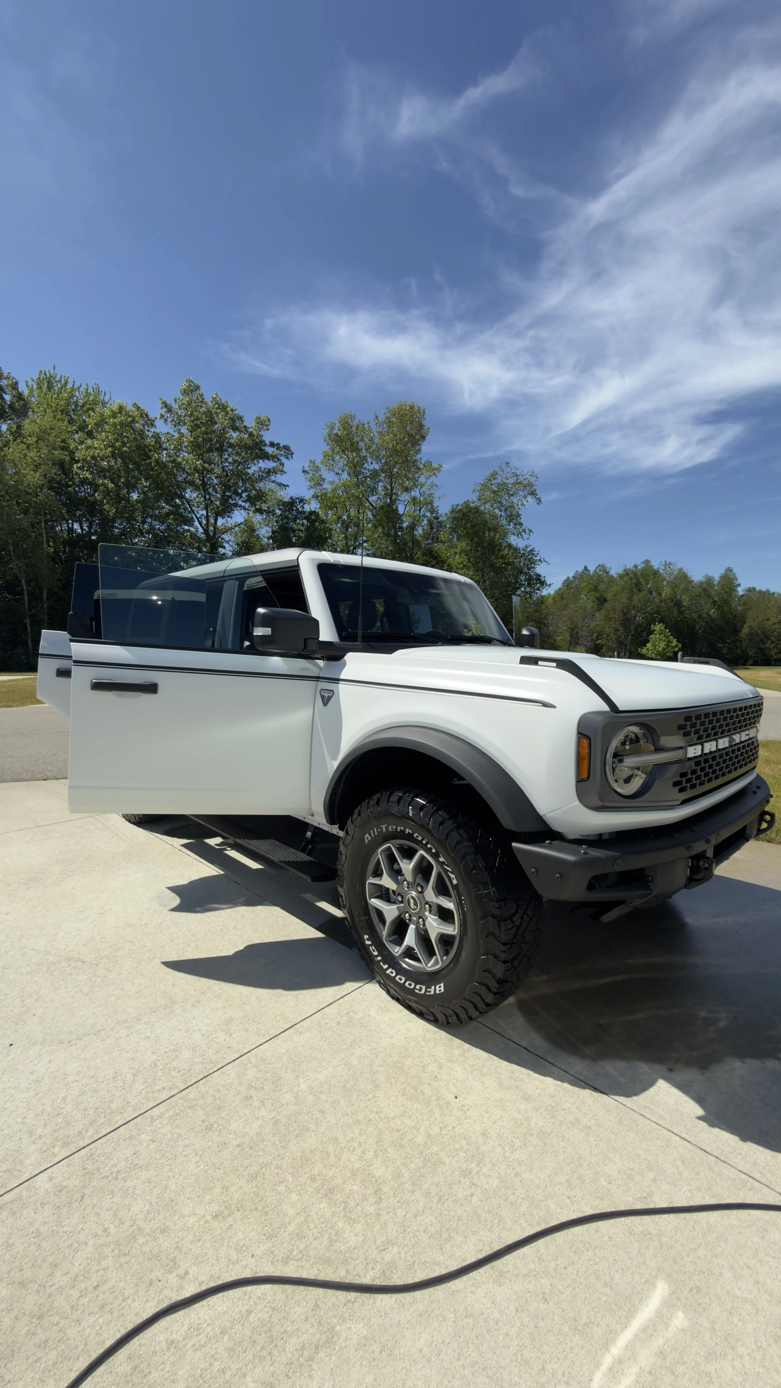 White off-road pickup truck parked on driveway under a blue sky with scattered clouds, surrounded by green trees.
