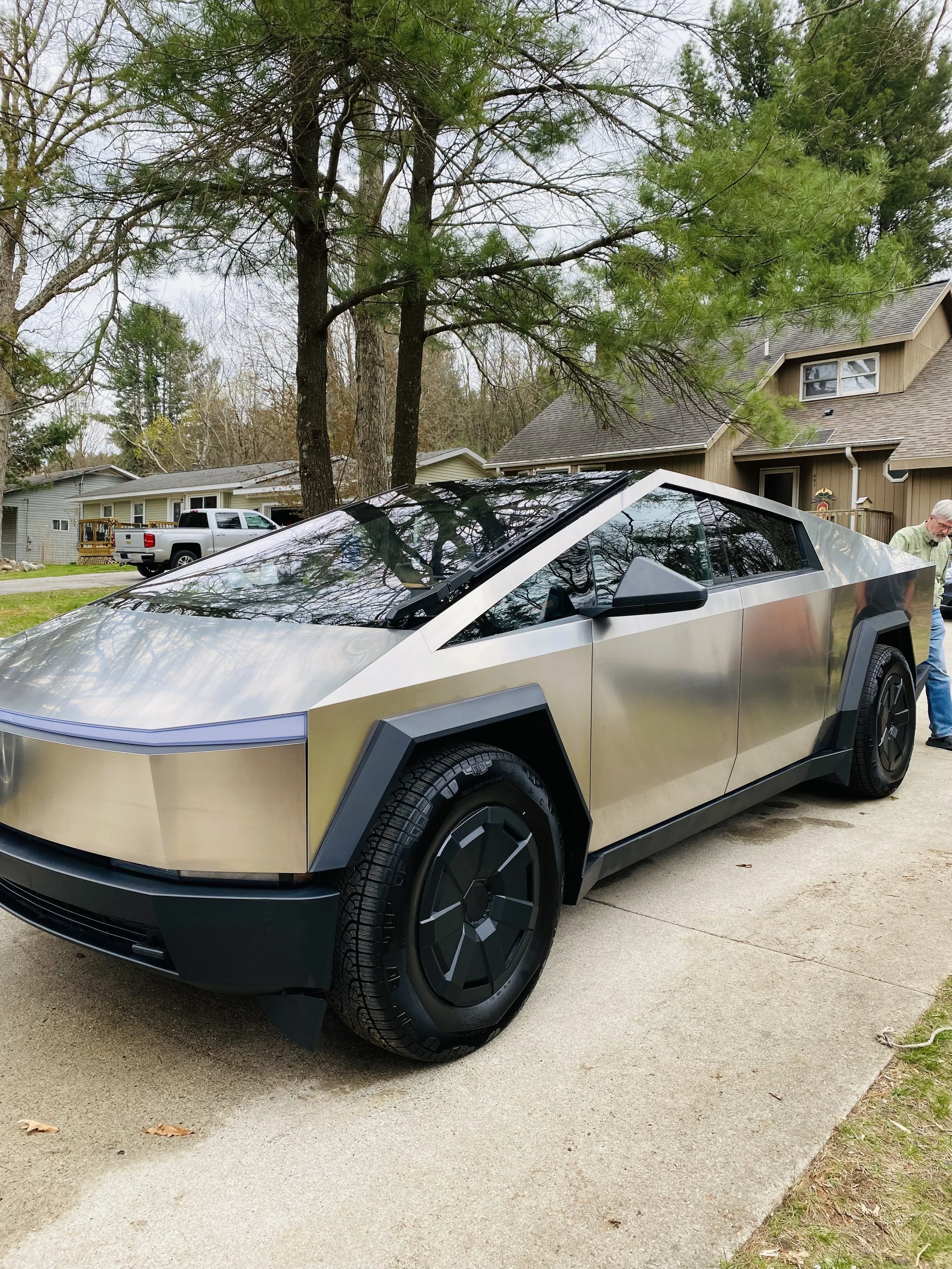 A silver futuristic-looking car parked on a driveway with trees and houses in the background.