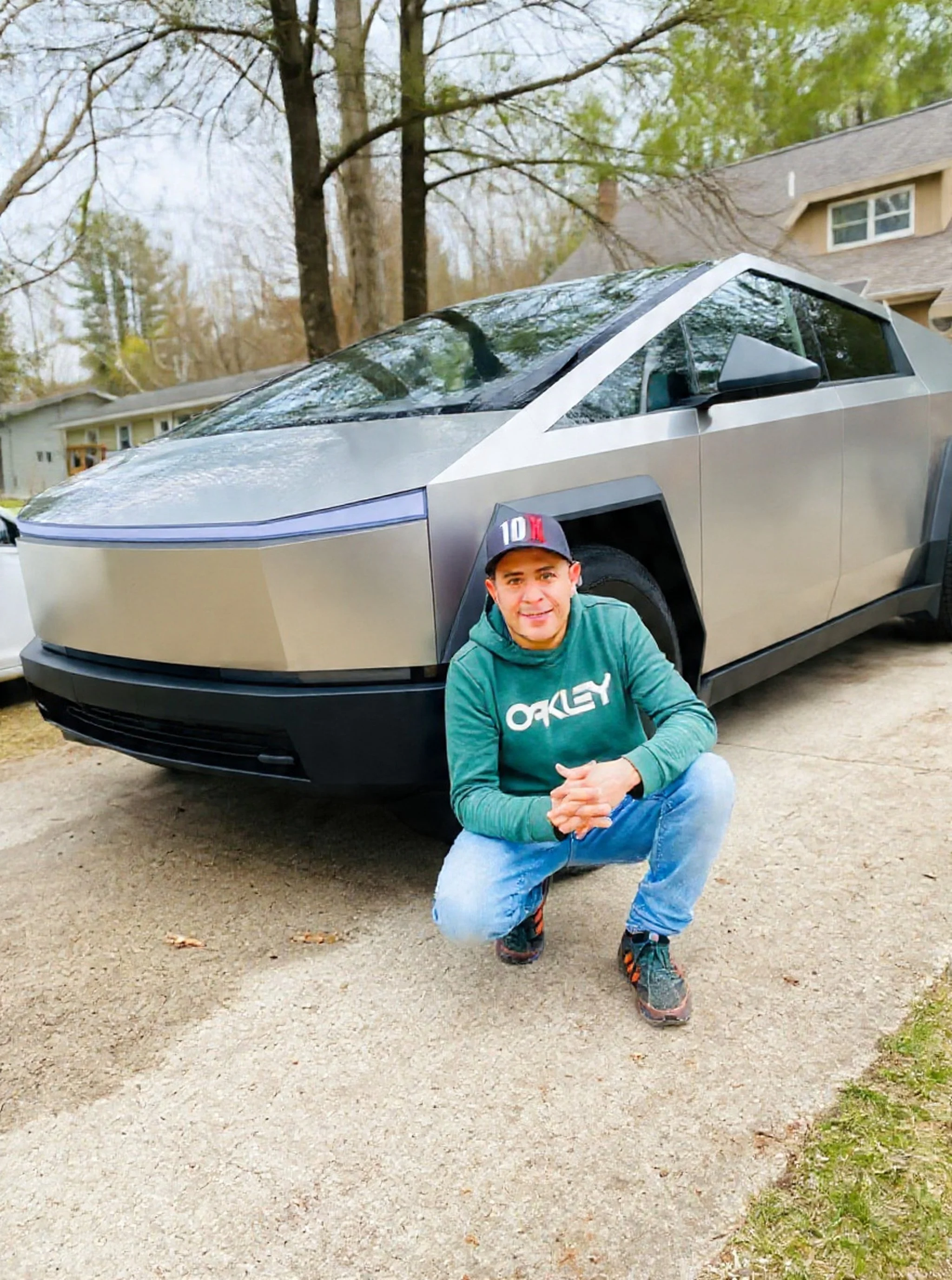 A man wearing a green Oakley hoodie and a black cap squatting in front of a futuristic silver electric vehicle, on a dirt driveway in front of suburban houses surrounded by trees.