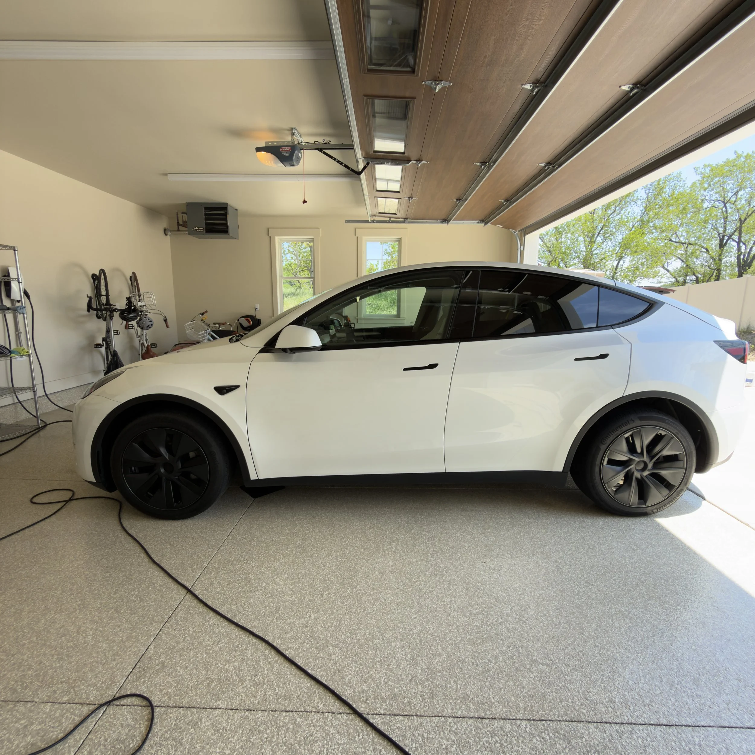 Inside a garage showing a white Tesla, bicycle racks with bikes, a garage door opener, and windows with trees outside.