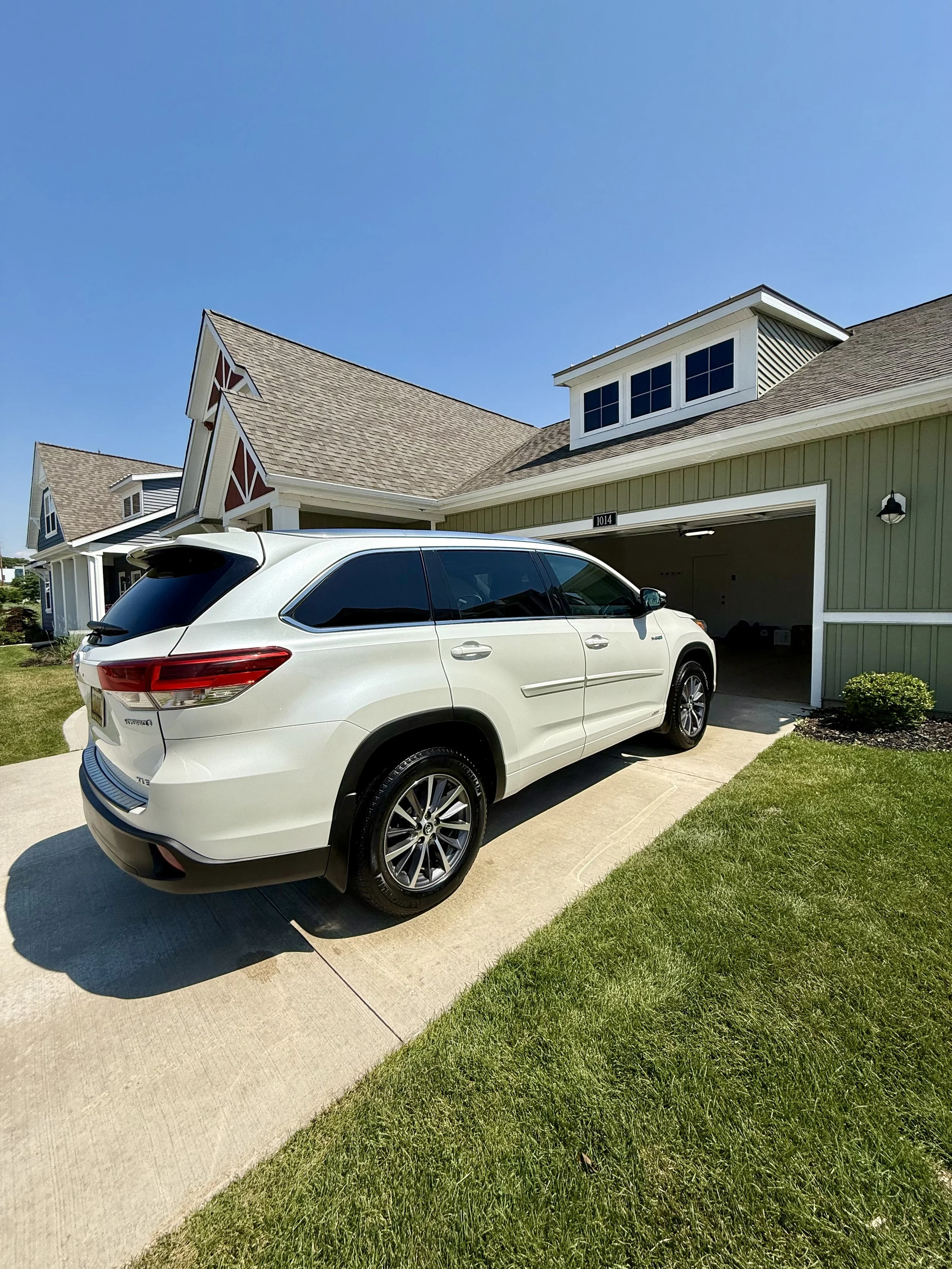 White SUV parked in driveway in front of a green house with a garage. The house has a gabled roof, white trim, and a small windowed dormer. The yard is grassy, and the sky is clear and blue.