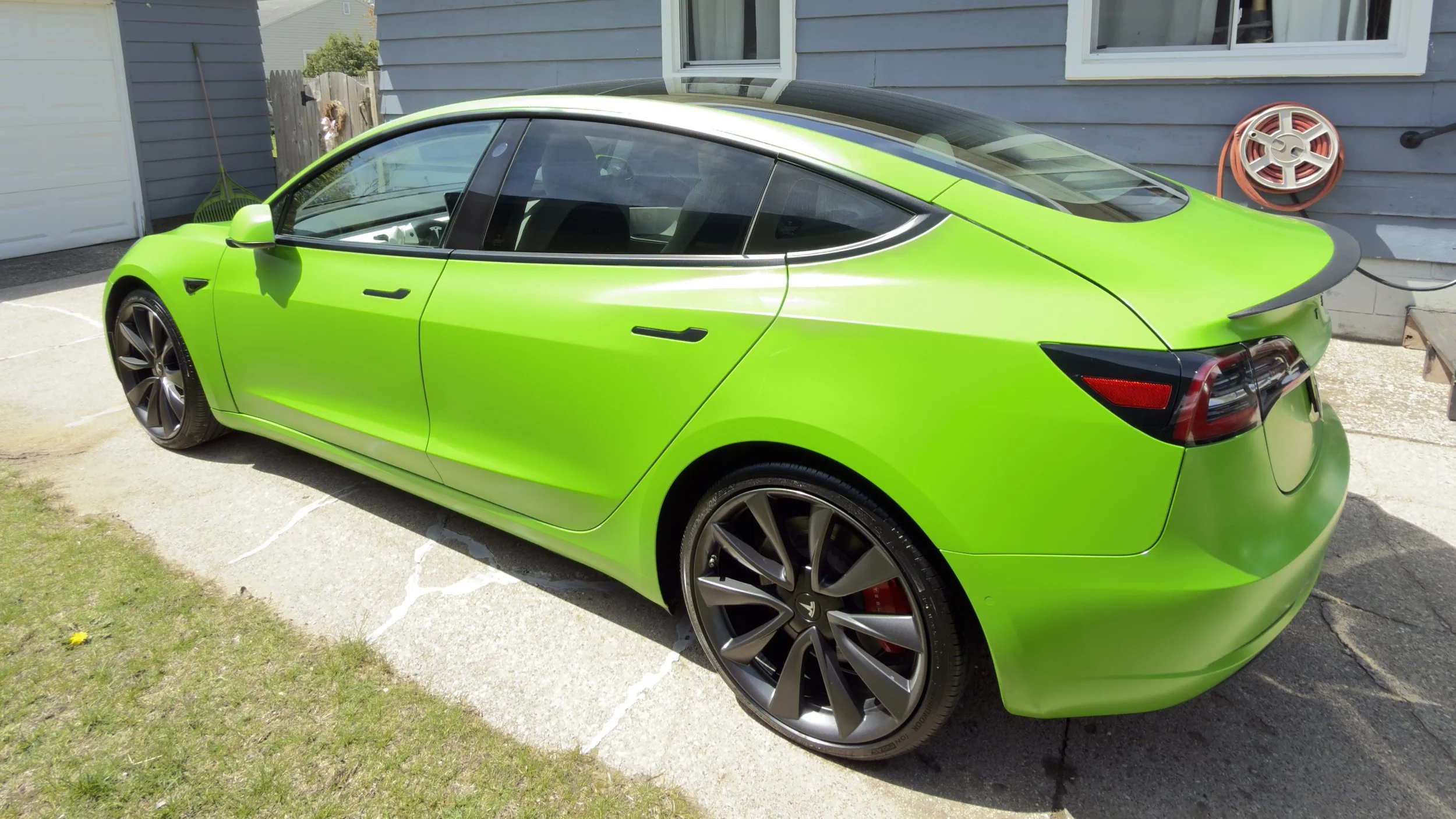 A bright green Tesla Model 3 electric car parked on a driveway beside a gray house with a window, a garden hose reel on the wall, and a patch of grass with a yellow dandelion flower.