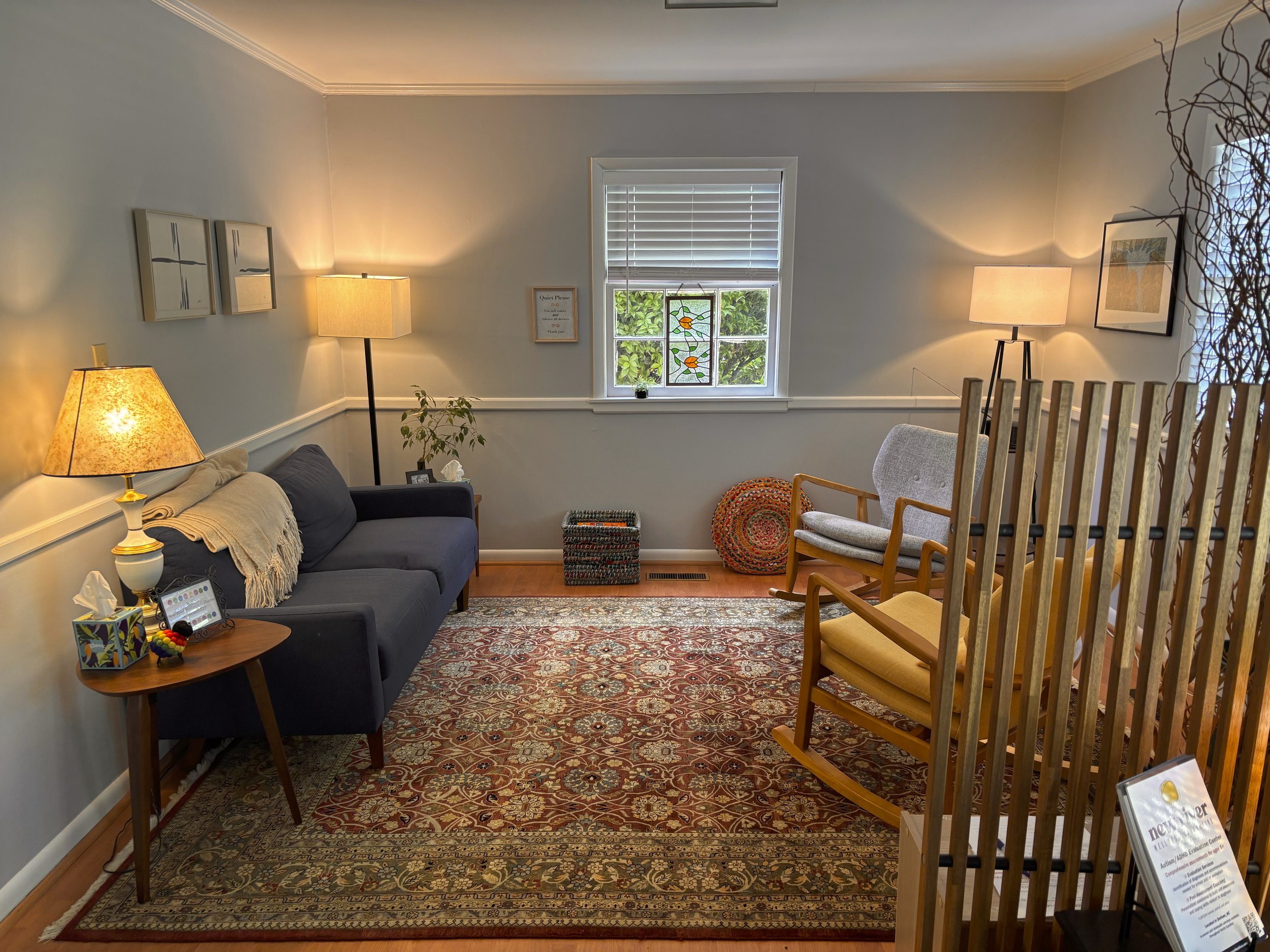 A cozy living room with a gray couch, a wooden side table, and various lamps. The room has a window with blinds, framed artwork on the walls, and a patterned rug on the wooden floor.