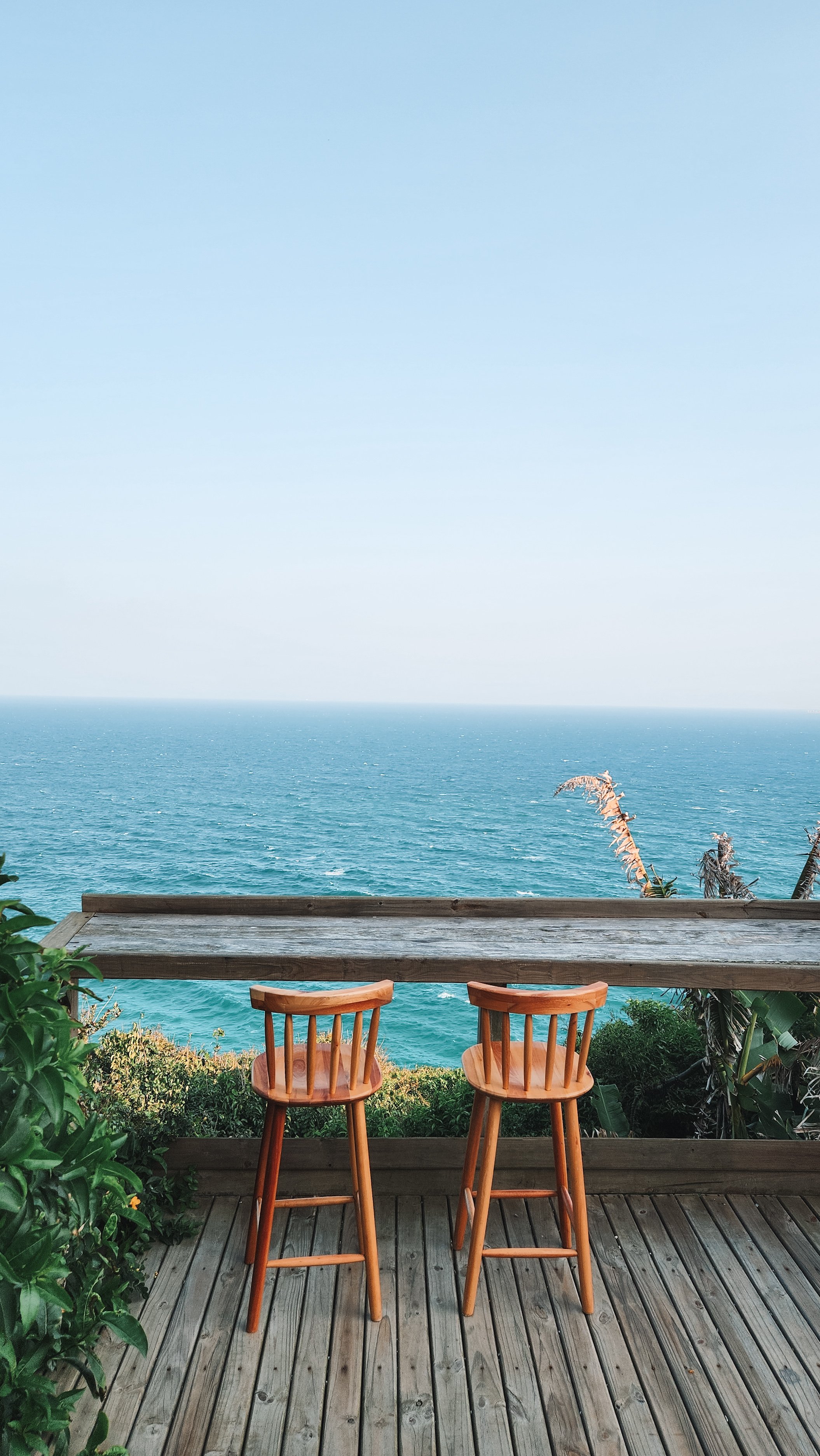 Two wooden chairs on a balcony overlooking the ocean with a blue sky, green foliage on sides, and a wooden railing in the foreground.