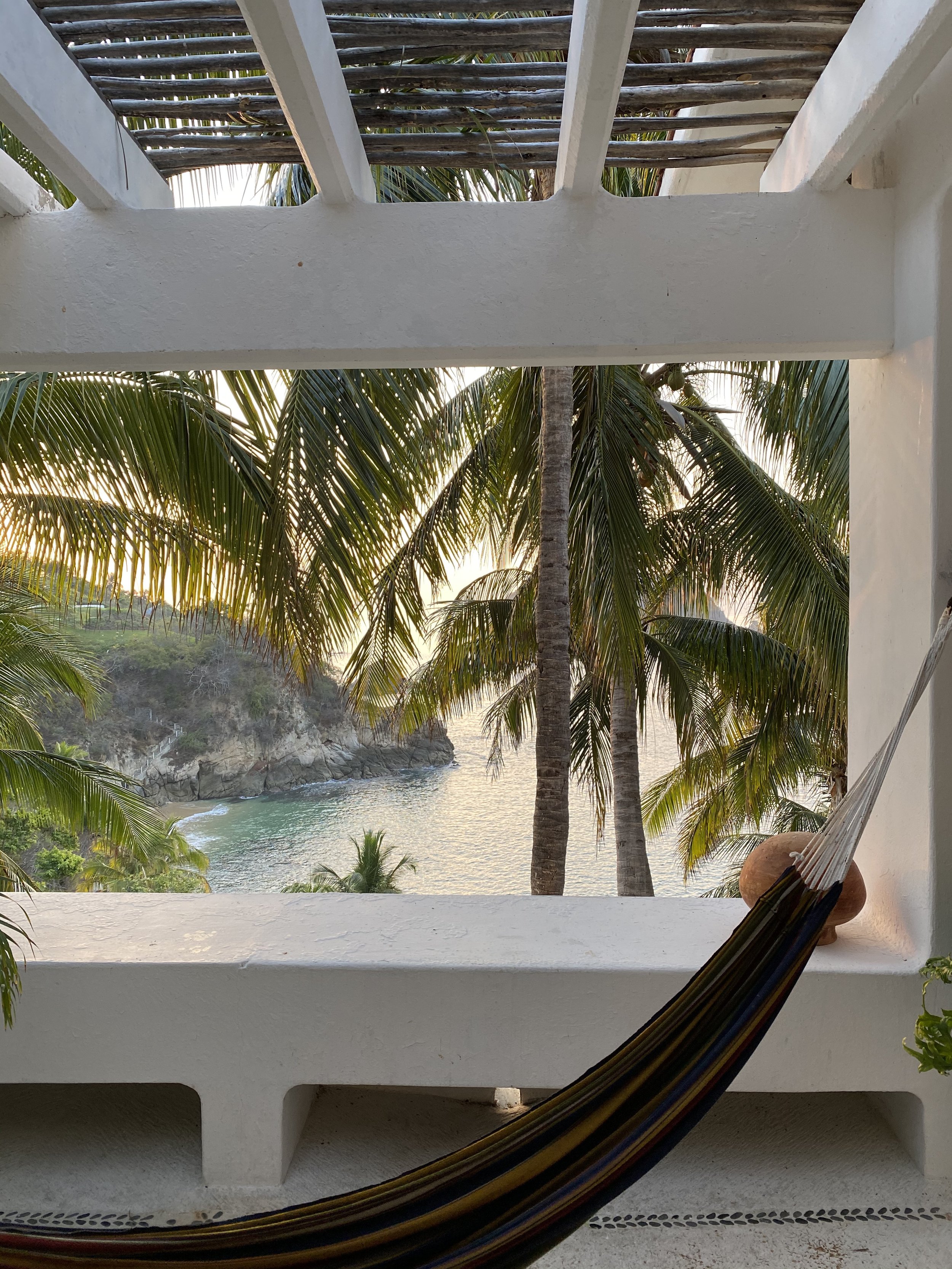View of palm trees and coastline seen through a window with a white wall and a hammock in the foreground.