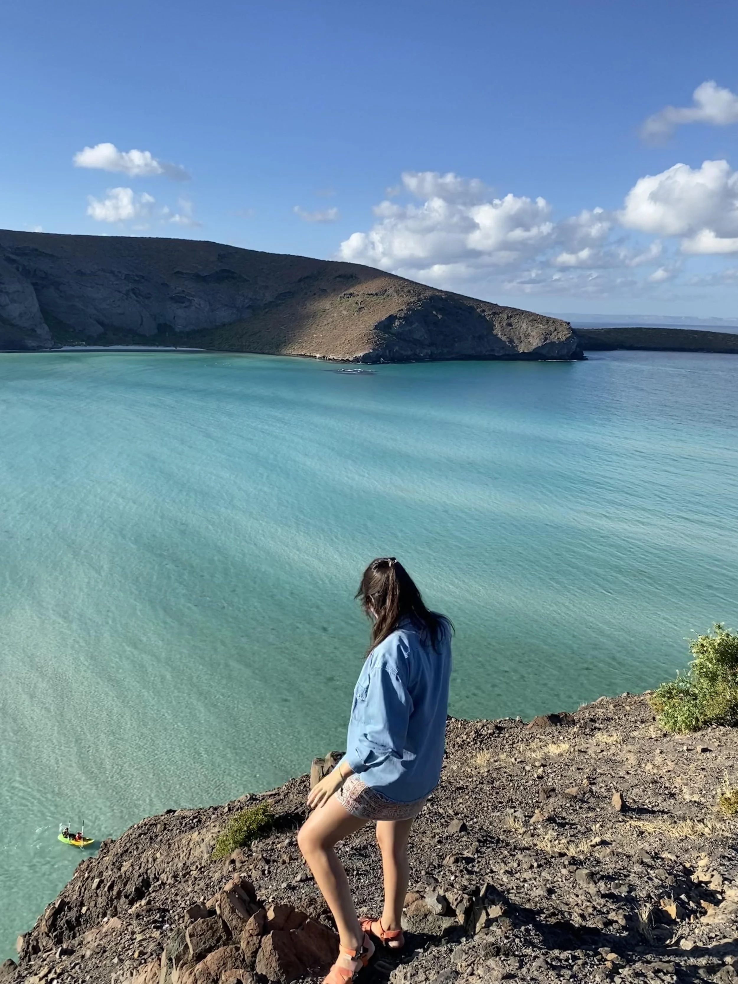 A person sits on rocks at a coastline with turquoise water and green hills in the background. The person is wearing a blue jacket, patterned shorts, and sandals, and is looking down at the water.
