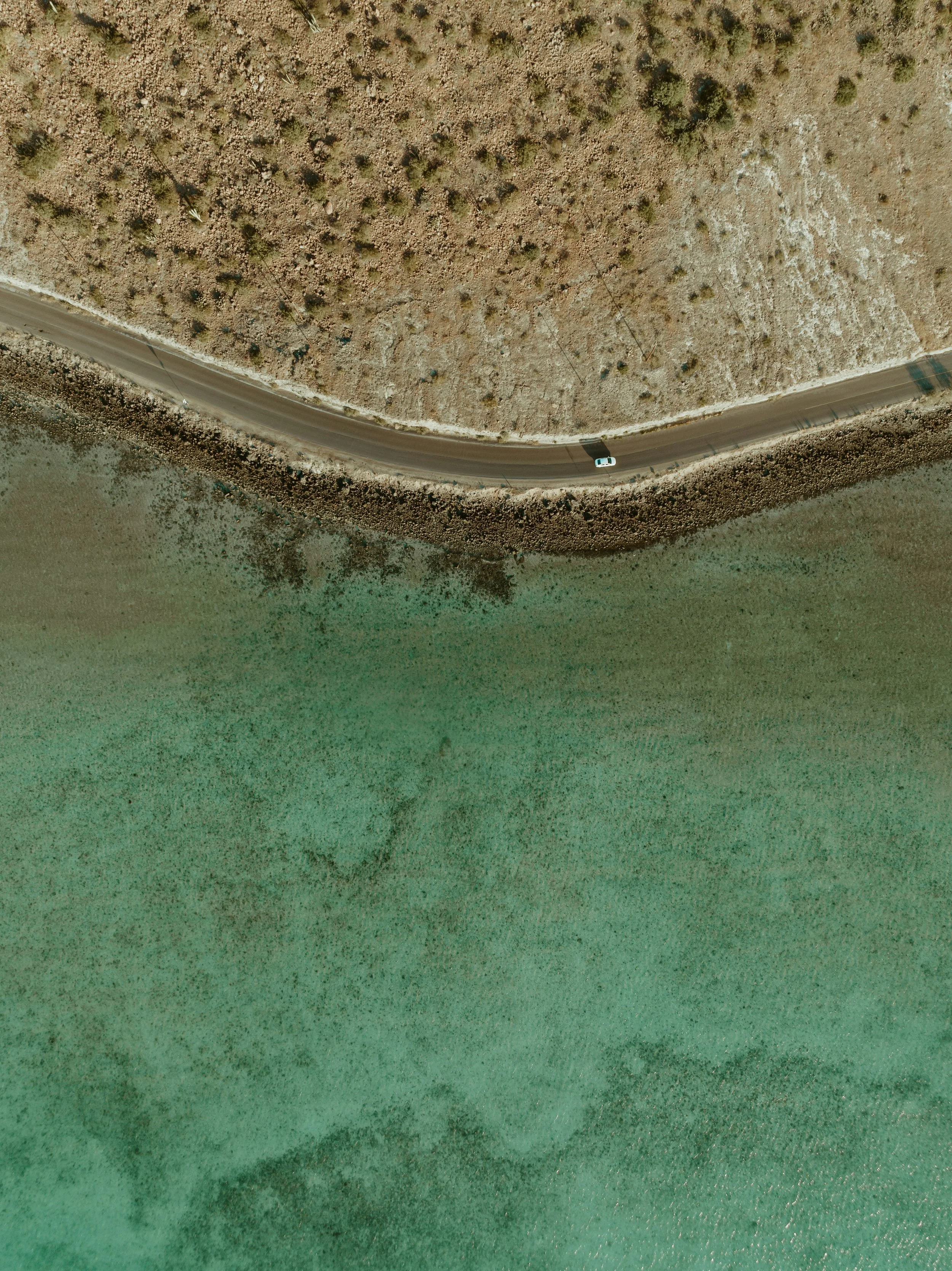 An aerial view of a winding road beside a body of water with arid land and sparse vegetation.