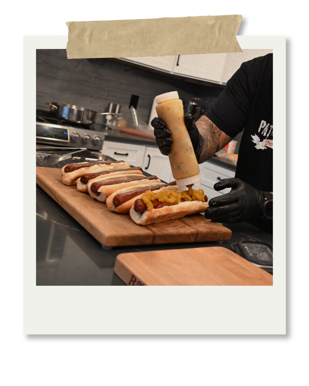 Chef preparing hot dogs with mustard and toppings on a wooden cutting board in a kitchen.