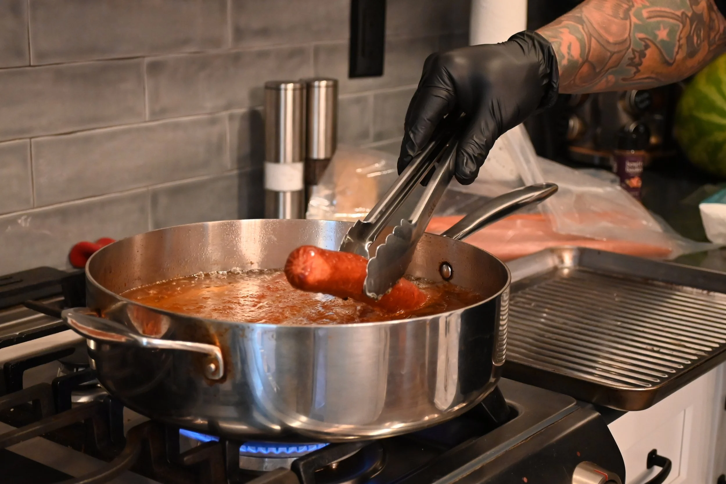 A person with tattooed arms wearing a black glove is using tongs to cook sausages in a skillet on a stovetop. The skillet contains a reddish sauce, and there are salt and pepper shakers on the countertop beside it.