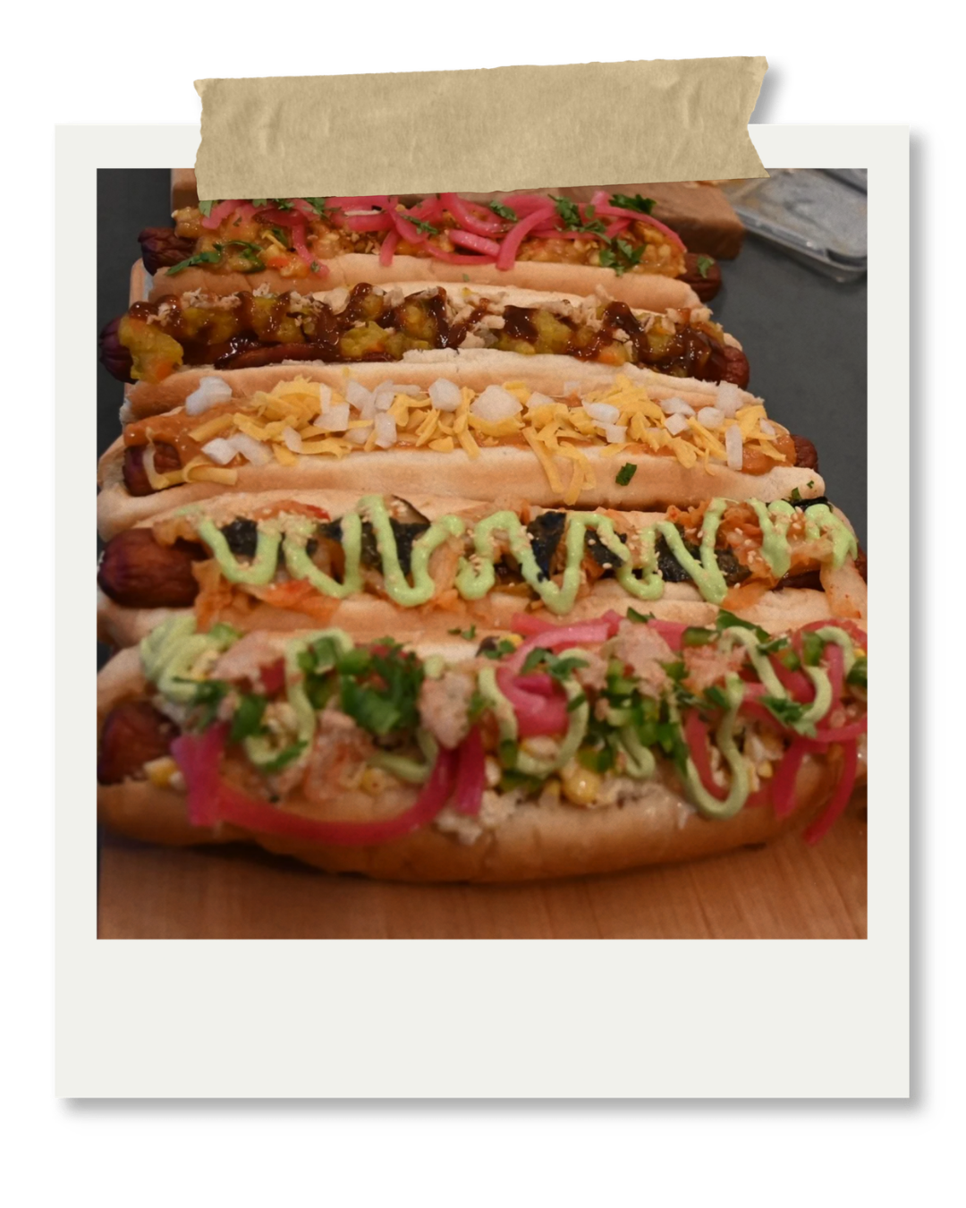 Five hot dogs with various toppings, including relish, chopped onions, mustard, cheese, and sauce, arranged on a wooden platter.