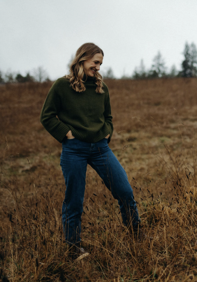 A woman standing in a grassy field with her hands in her pockets, wearing a green sweater and blue jeans, smiling and looking down.