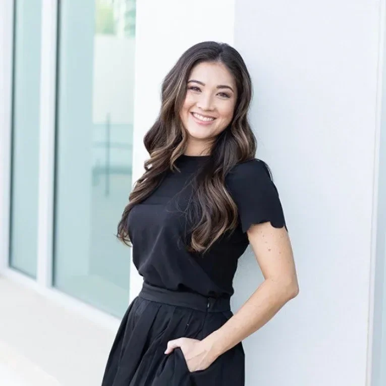 A young woman with long, wavy brown hair is smiling and standing against a white wall, wearing a black t-shirt and black skirt.