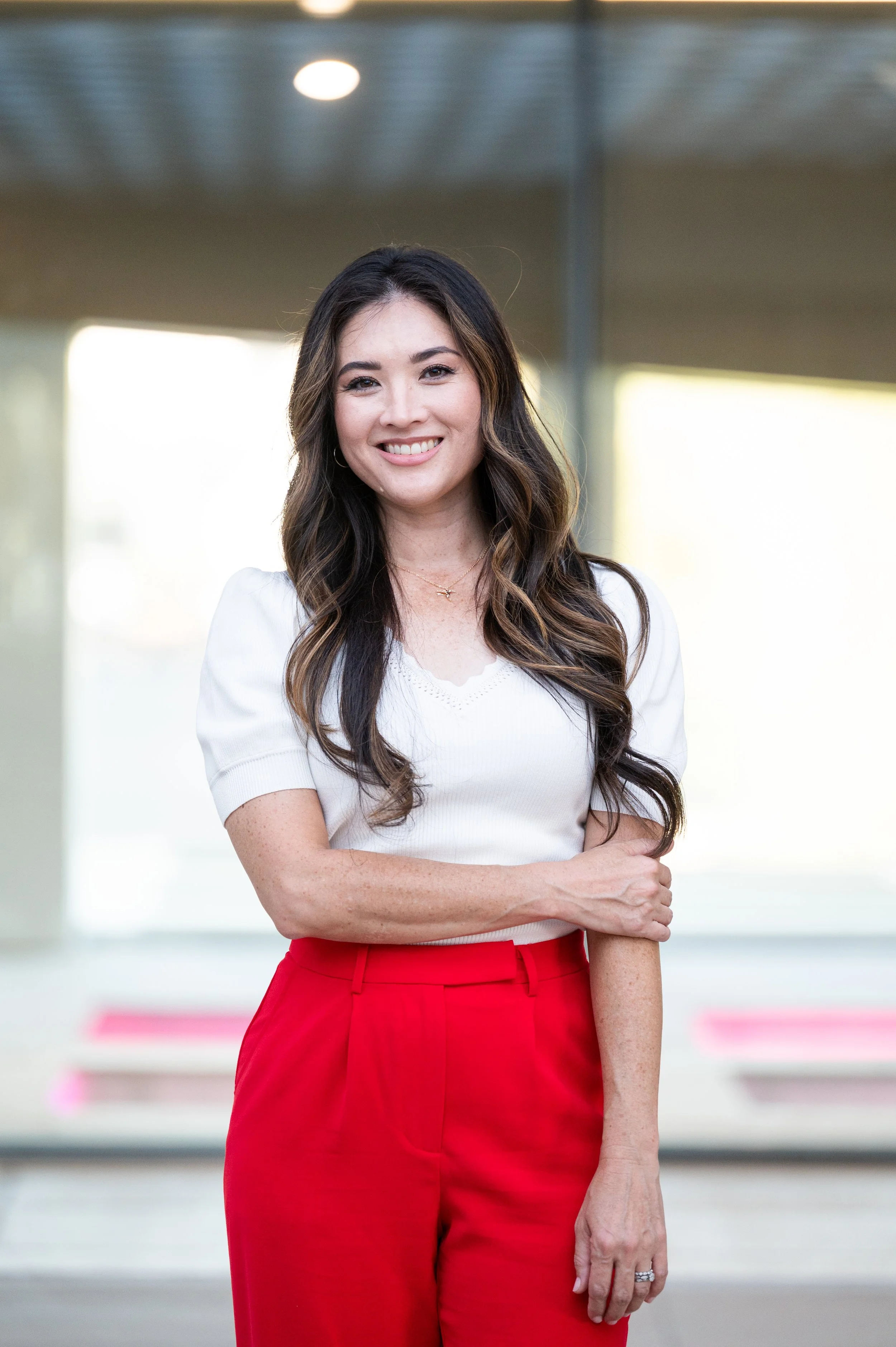 A woman with long dark hair and a bright smile, wearing a white top and red pants, standing in an indoor corridor with a blurred background.