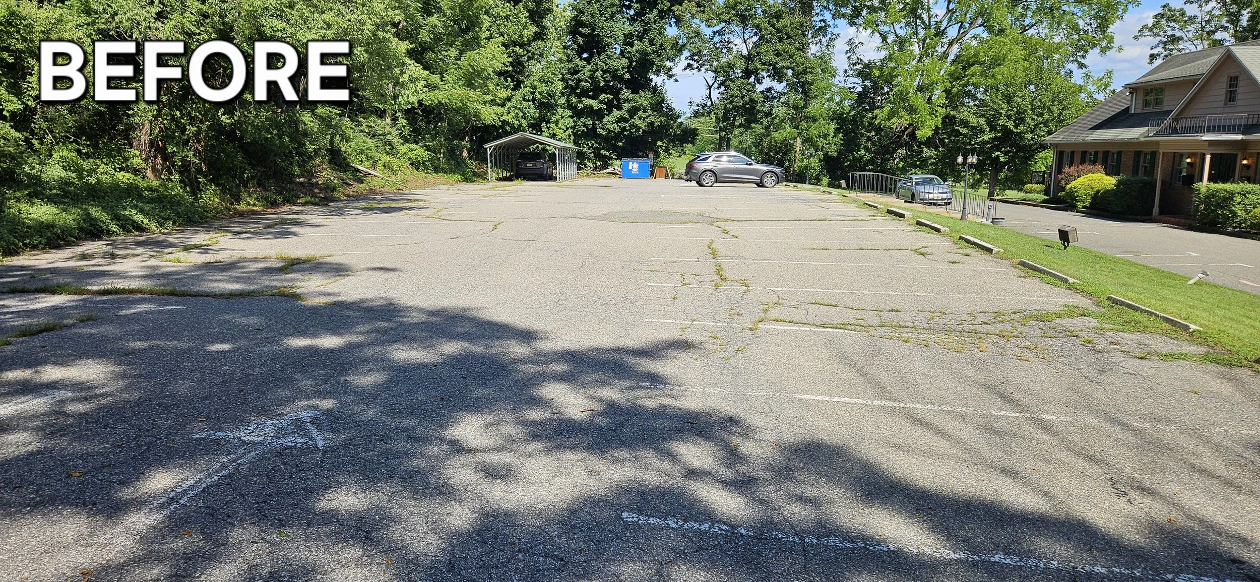 Empty parking lot with cracked asphalt, surrounded by trees and a building on the right.