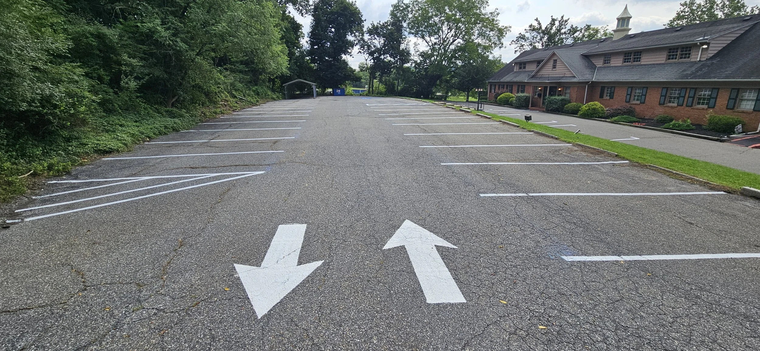 Empty parking lot with white arrows painted on the asphalt, surrounded by trees and a brick building with a porch.