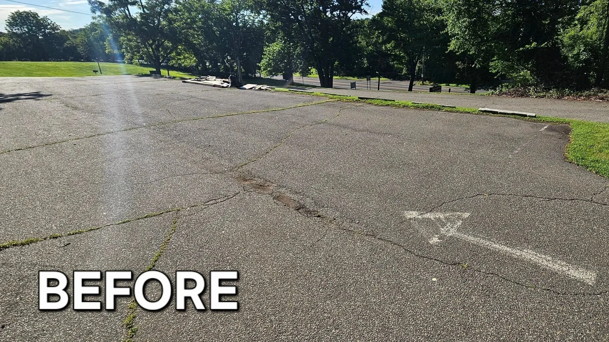 A cracked asphalt parking lot with patches of grass growing through the cracks, surrounded by green trees and a grassy area in the background, under a partly cloudy sky.