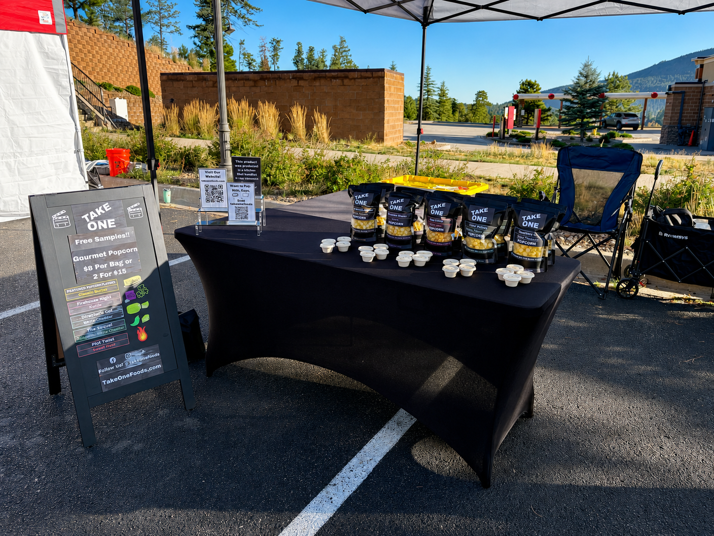 Outdoor market stall with a black tablecloth offering free samples of gourmet popcorn, featuring bags labeled 'Take One' and small cups for tasting, with a sign advertising popcorn for $8 per bag or 2 for $15.