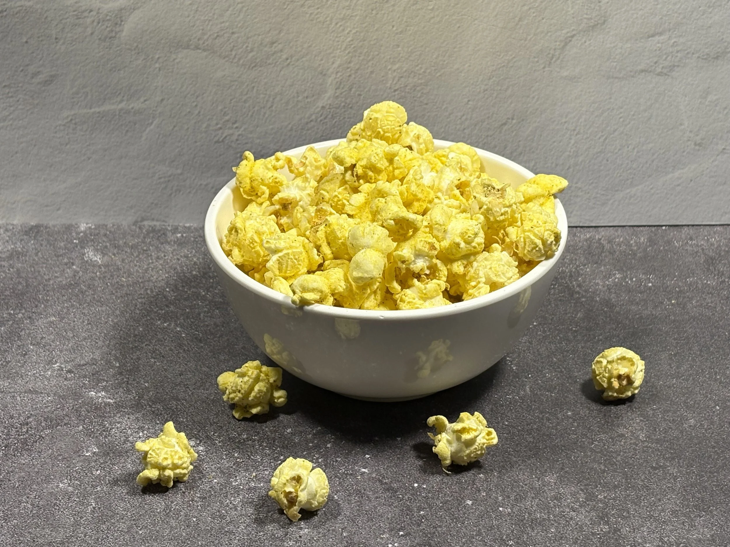 A white bowl filled with yellow popcorn on a gray countertop, with some popcorn pieces scattered around the bowl.