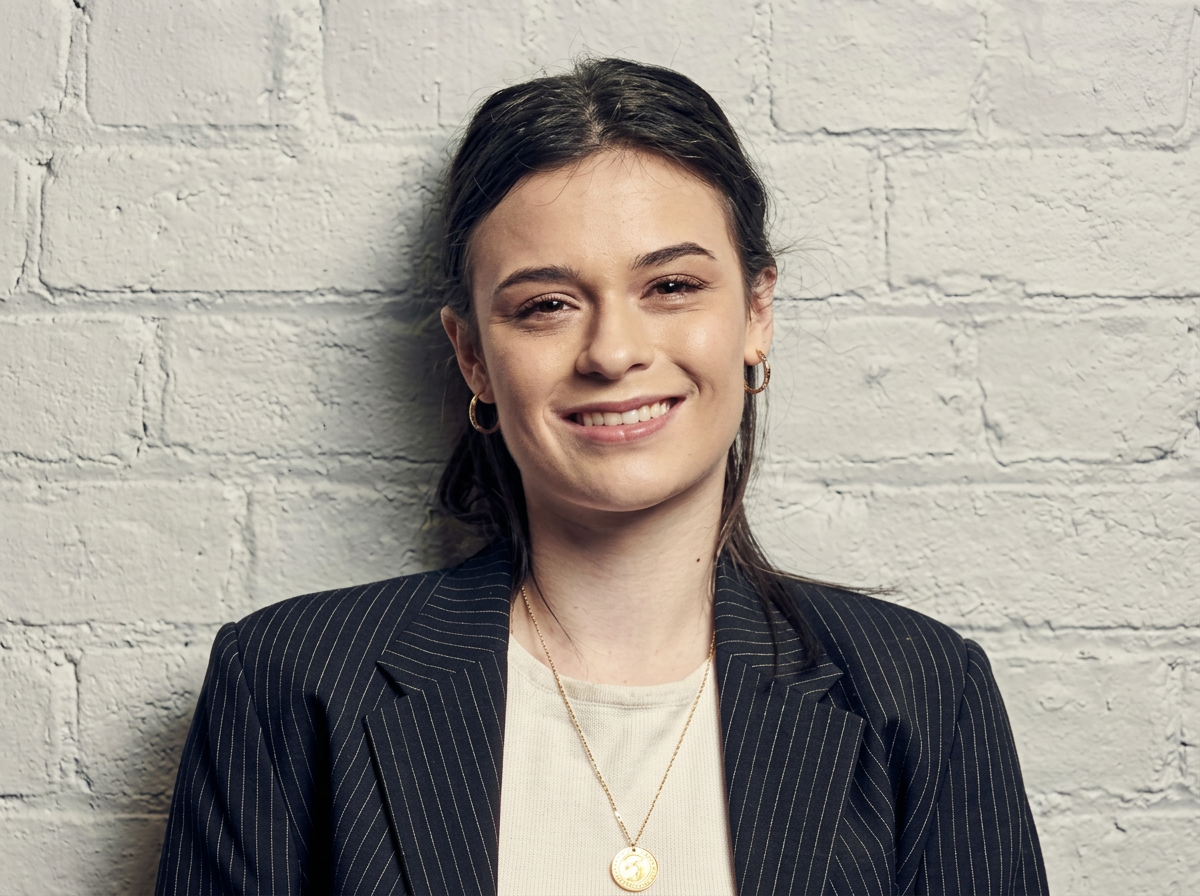 A young woman smiling, wearing a black pinstripe blazer, gold hoop earrings, a gold necklace with a pendant, and a white top, standing against a white brick wall.