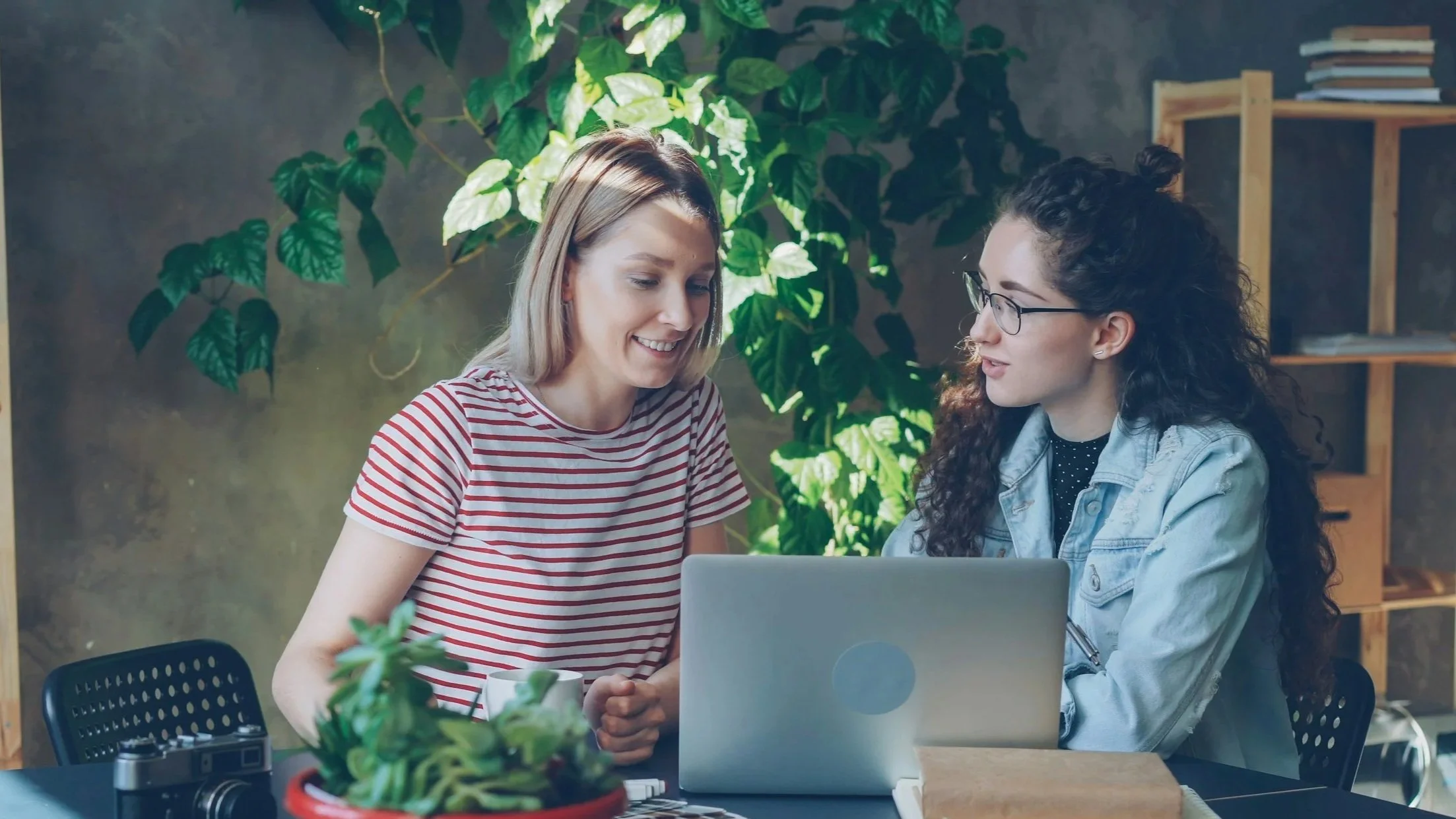 Two women sitting at a table with a laptop, looking at it together in a room with green plants and bookshelves.