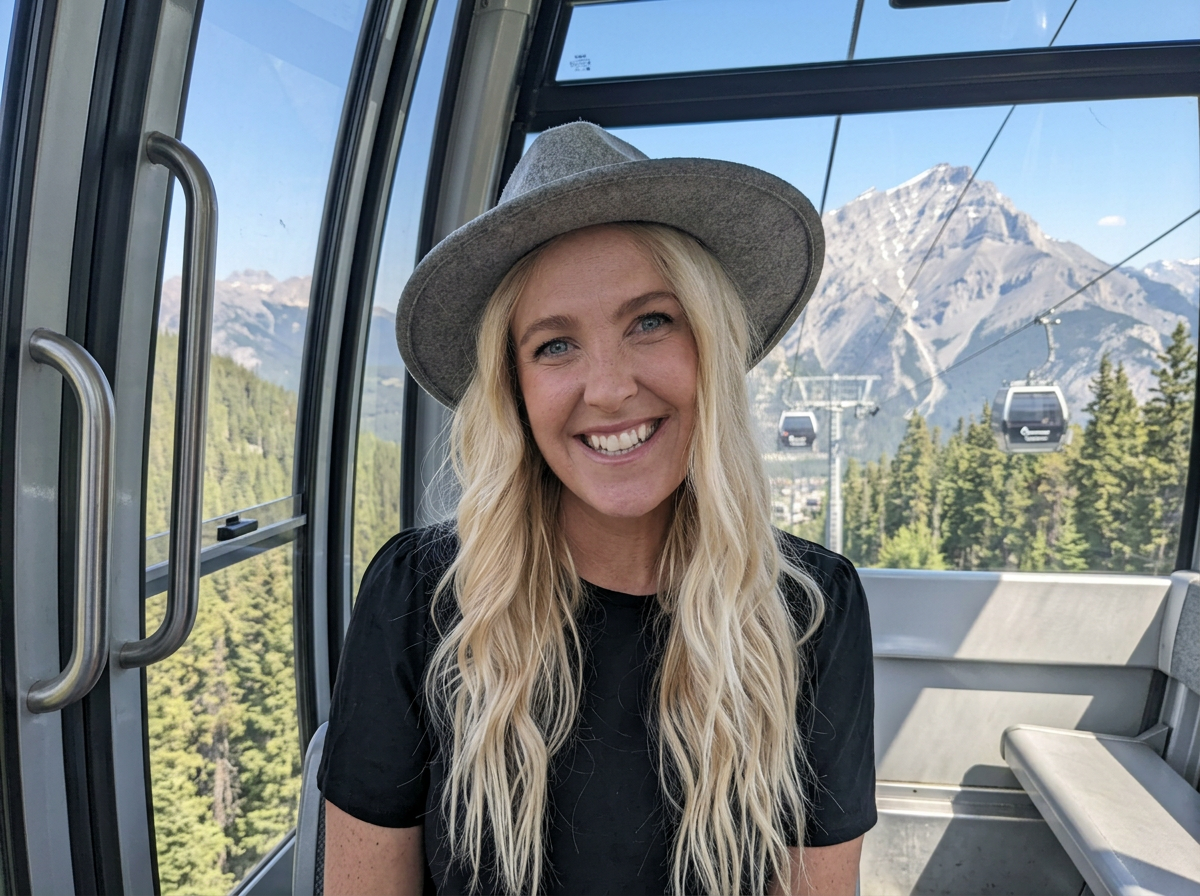 Woman with long blonde hair wearing a gray hat, smiling inside a gondola with mountain scenery and ski lifts in the background.