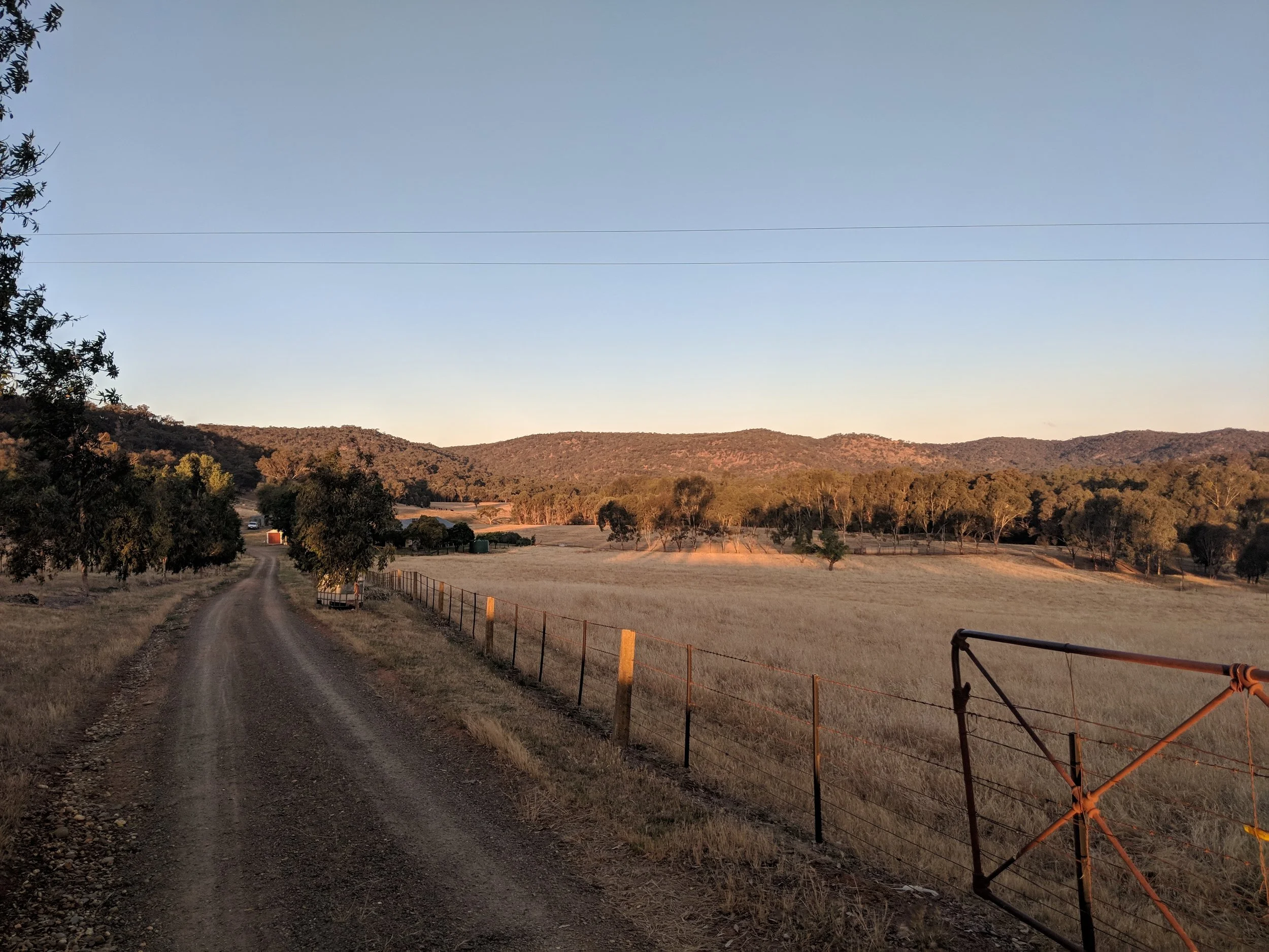 A rural landscape with a dirt road, dry grass, trees, rolling hills in the background, and a fallen fence gate on the right. The sky is clear with a few clouds.