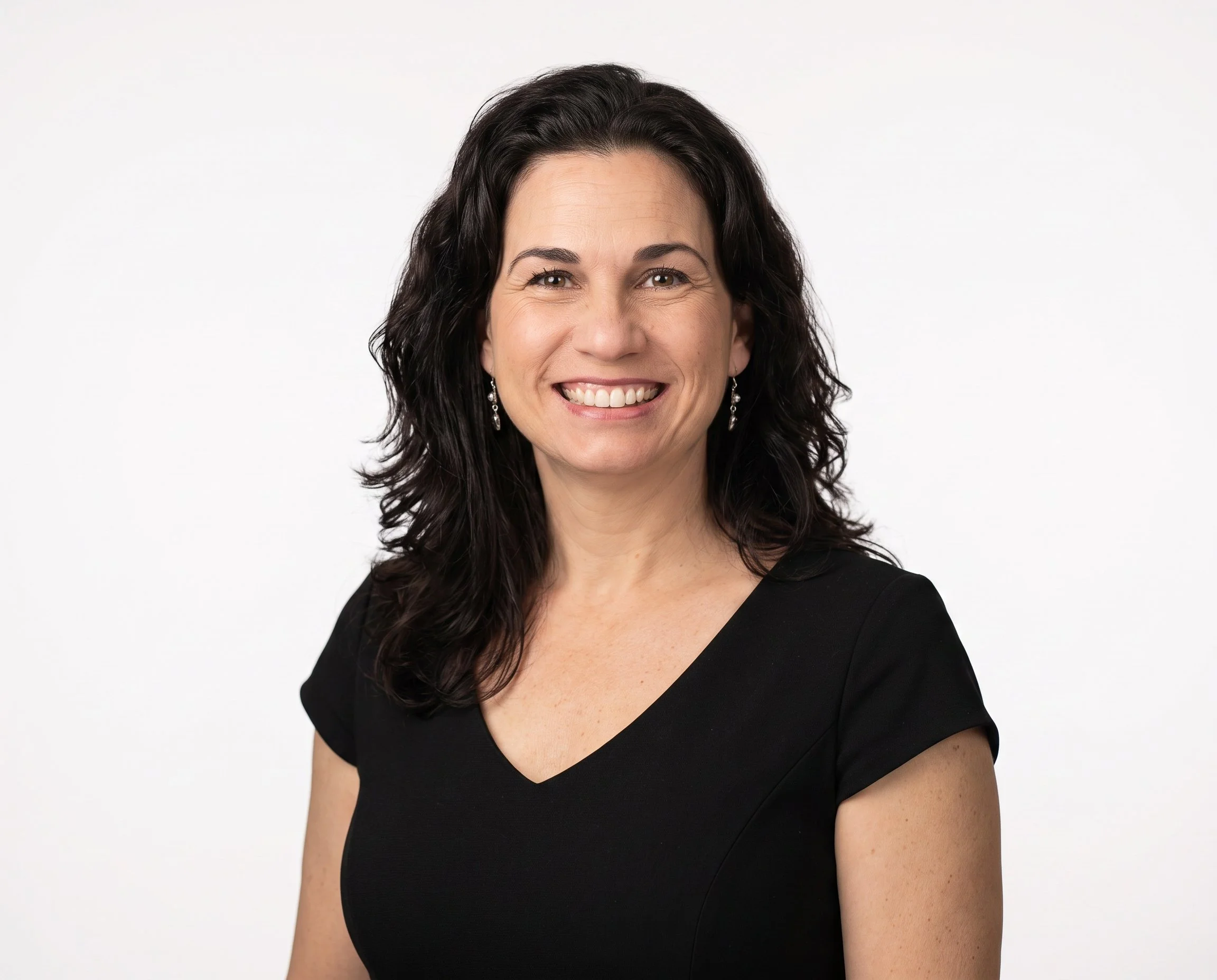 A woman with dark wavy hair smiling, wearing a black V-neck dress and earrings, against a plain white background.