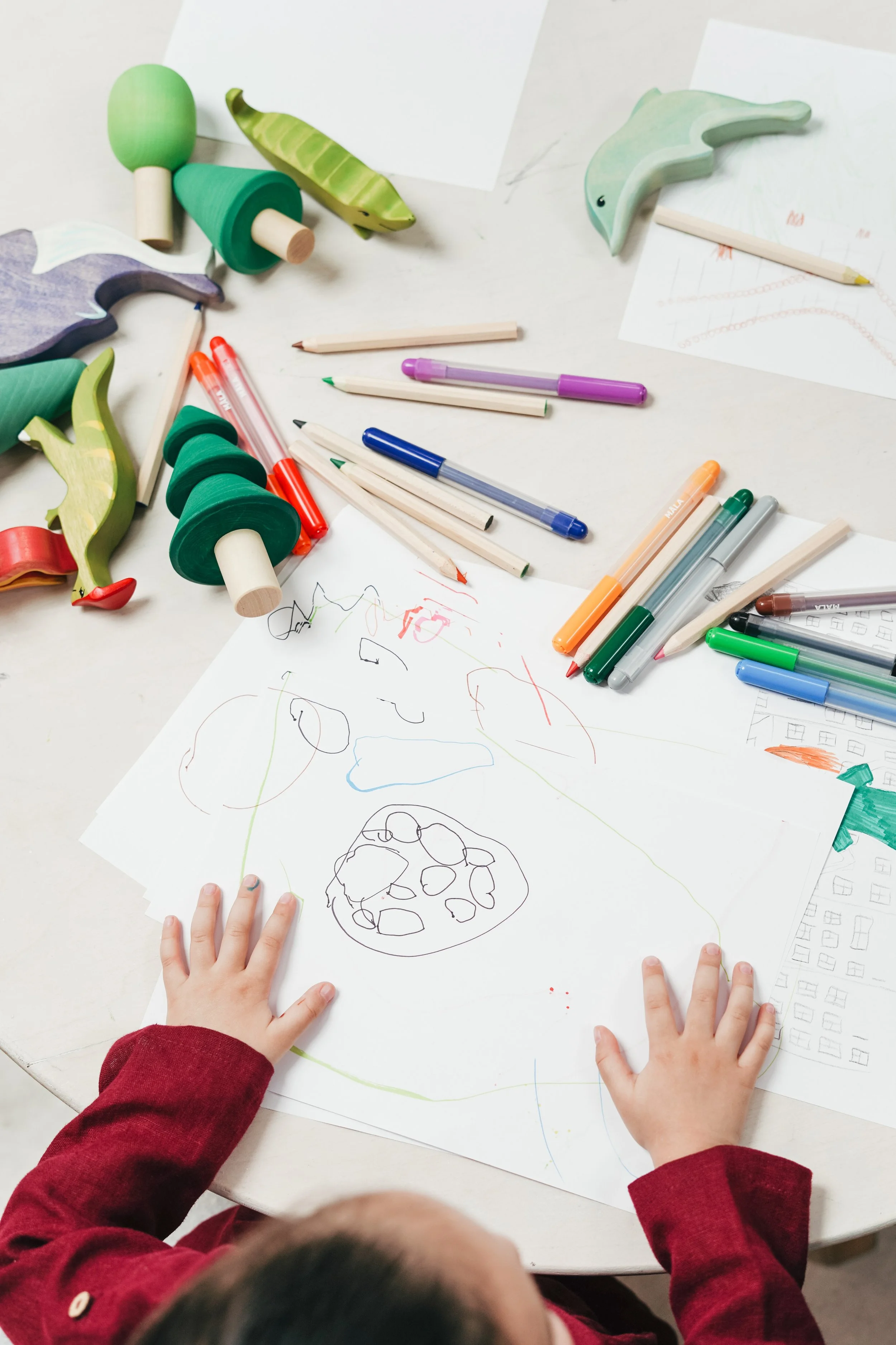 Child's hands drawing with colored and black pens on a large sheet of paper, surrounded by wooden animal-shaped toys and paper with sketches.