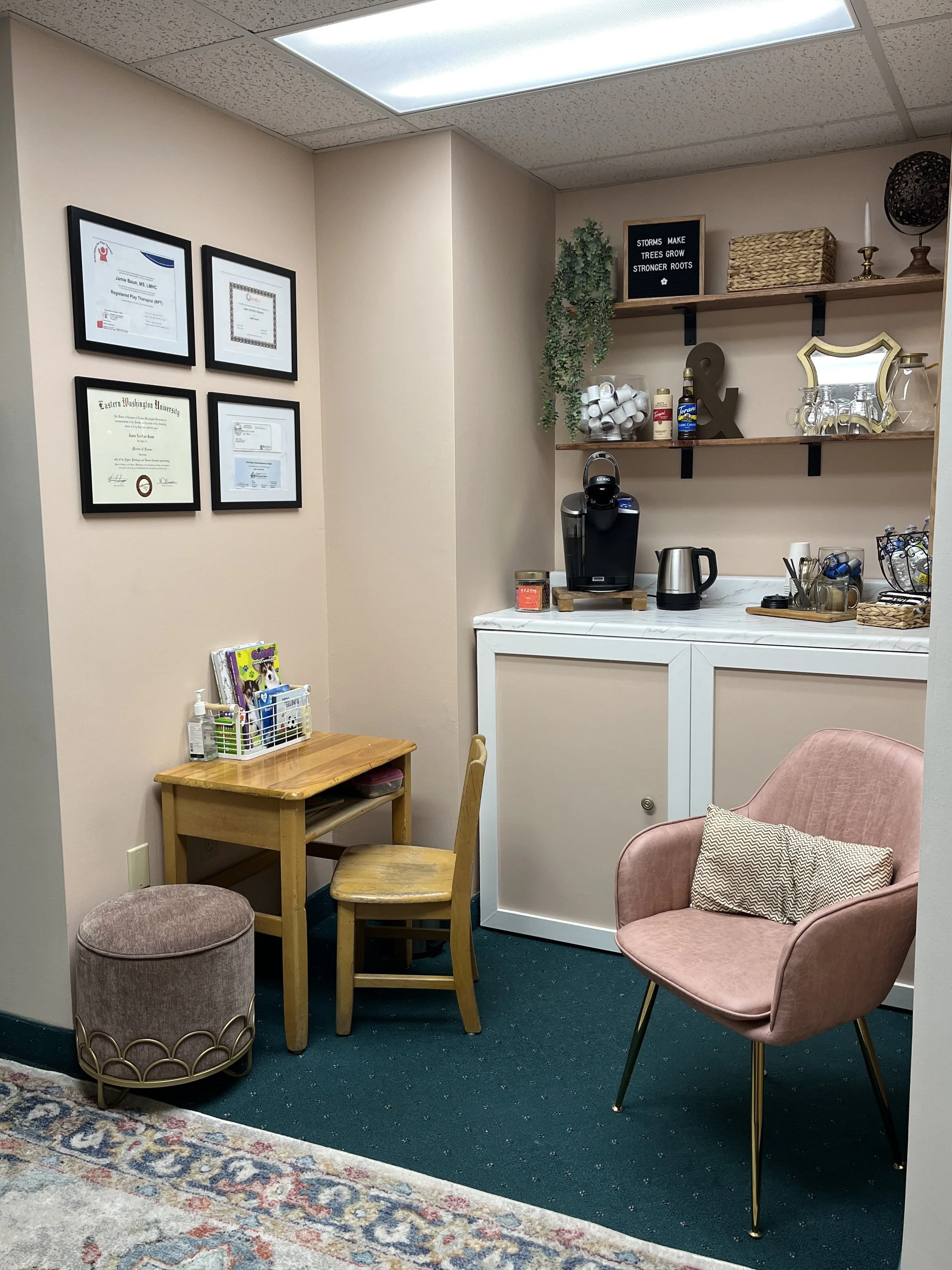 The room features a beige wall with four framed certificates, a small wooden table with a chair, a pink armchair with a patterned pillow, a brown ottoman, and a white cabinet with a marble top. On the marble counter, there is a Keurig coffee machine, an electric kettle, cups, and some office supplies. Two wooden shelves on the wall hold decorative items including a small plant, a black letter board with the message "STORMS MAKE TREES GROW STRONGER ROOTS," a woven basket, candles, and glassware.
