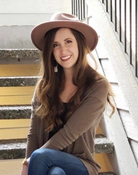 Young woman with long wavy hair wearing a pink hat, brown cardigan, and jeans, sitting on outdoor stairs and smiling at the camera.