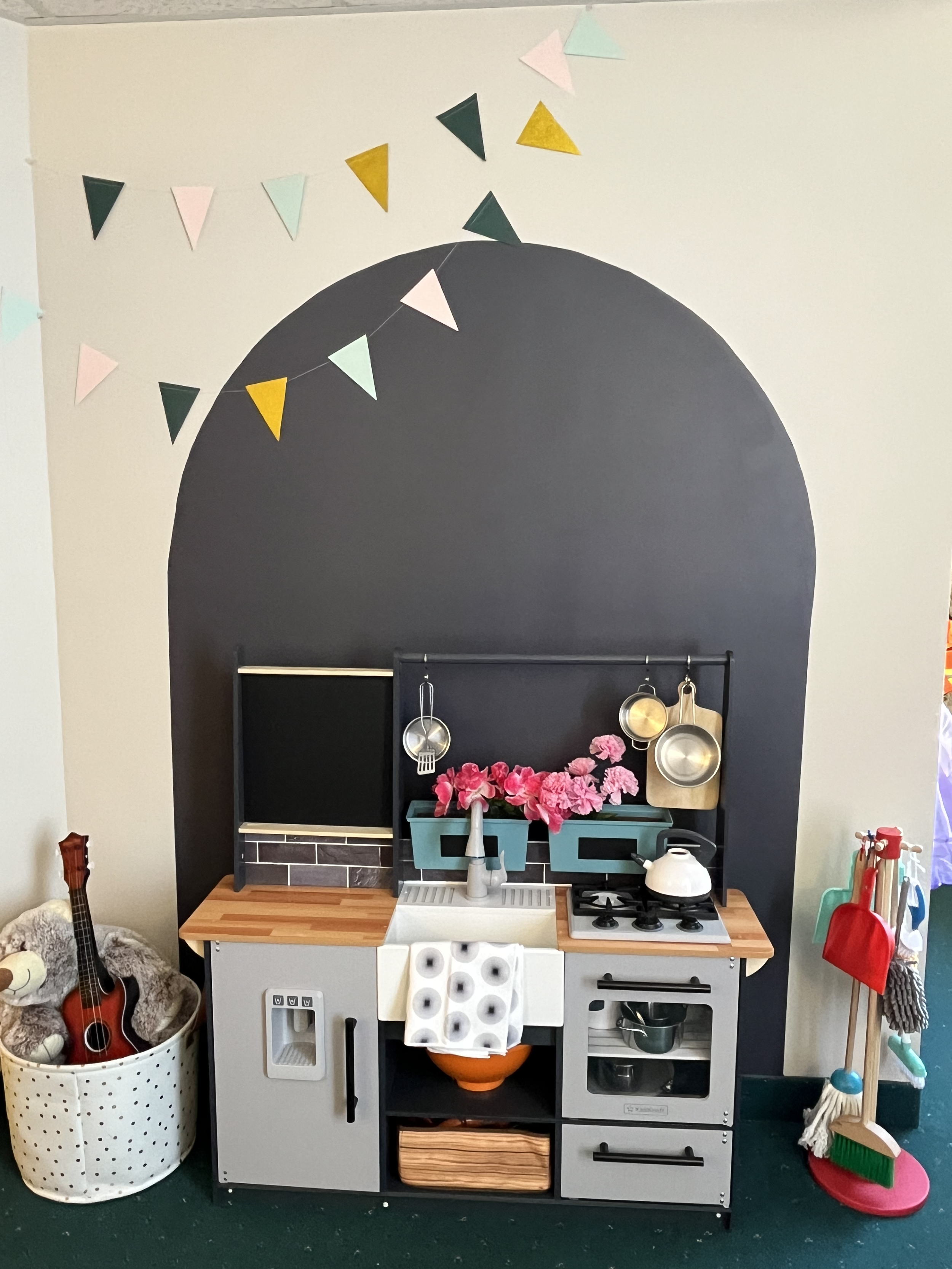 A children's play kitchen with a sink, stove, and storage compartments. There are pink flowers in a teal container, kitchen utensils hanging, and a small blackboard. To the left, a stuffed bear and a small guitar are in a white basket. To the right, cleaning brushes are on a stand. The background wall has pastel-colored triangular flags hanging above the play kitchen.
