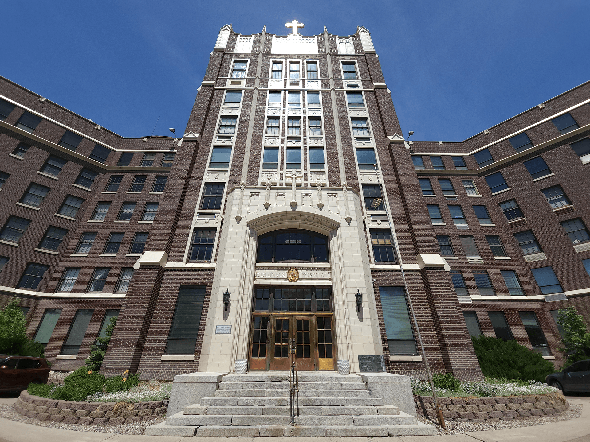 Front view of Columbus Hospital, a large brick building with an arched entrance and steps leading up to it, under a clear blue sky.