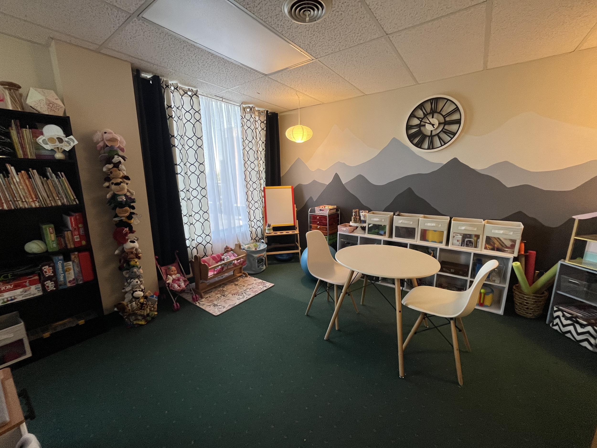 A colorful children's playroom with a mountain-themed wall mural, shelves filled with books and toys, a round table with white chairs, a cozy play area with stuffed animals, a clock on the wall, and patterned curtains.