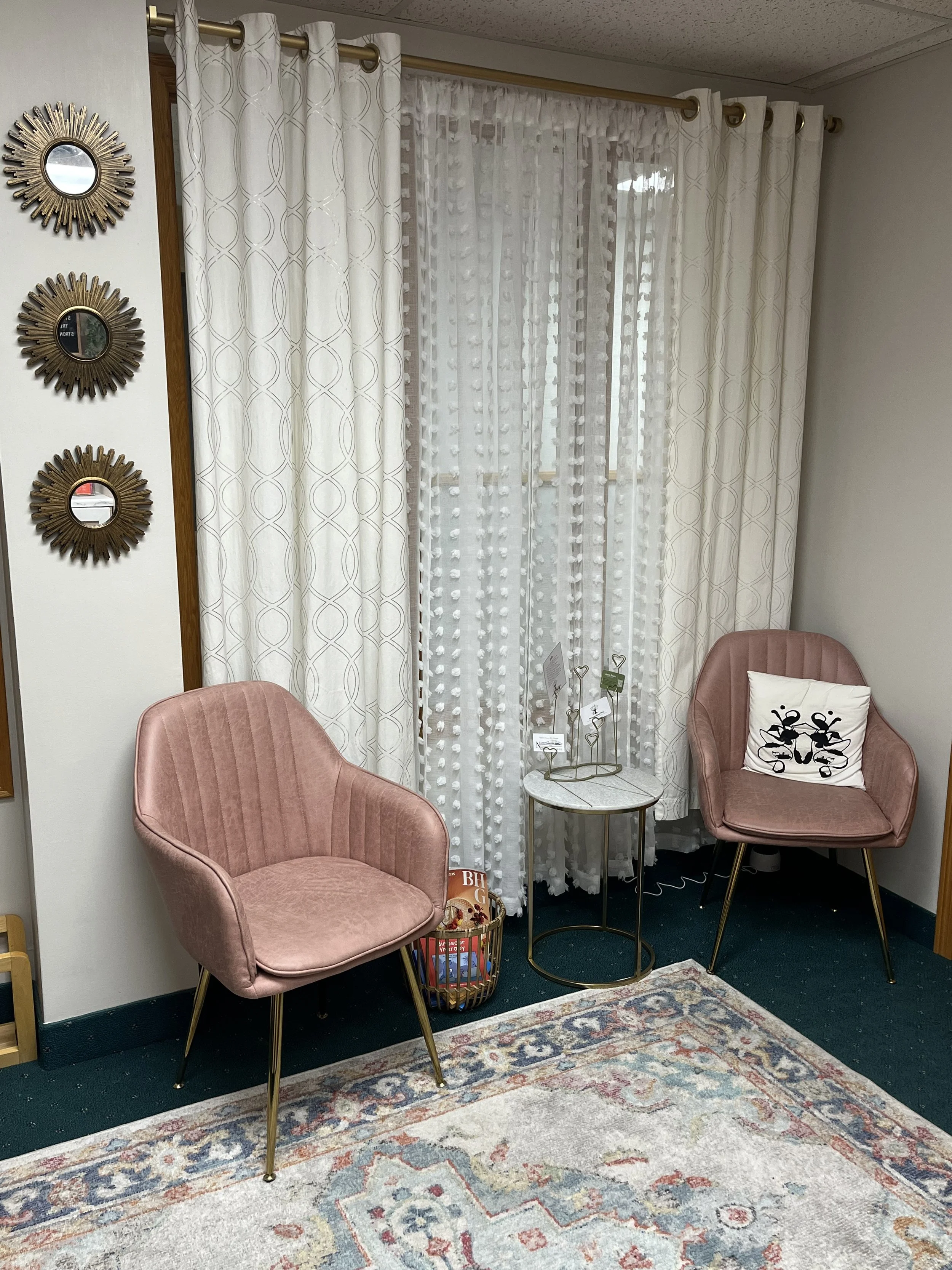 A cozy sitting area with two pink velvet armchairs, a small round table, and decorative elements such as a basket of books, three round mirrors with sunburst frames on the wall, and white curtains with gold rings covering a window. The floor is partially covered with a patterned rug.