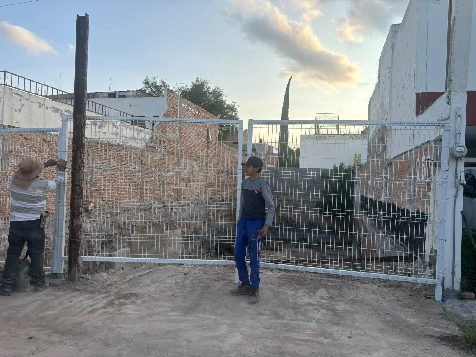 Dos personas, una de ellas ajustando una cerca metálica blanca y un joven parado frente a la cerca, en un entorno urbano con paredes de ladrillo y edificios en el fondo, durante el atardecer con cielo parcialmente nublado.