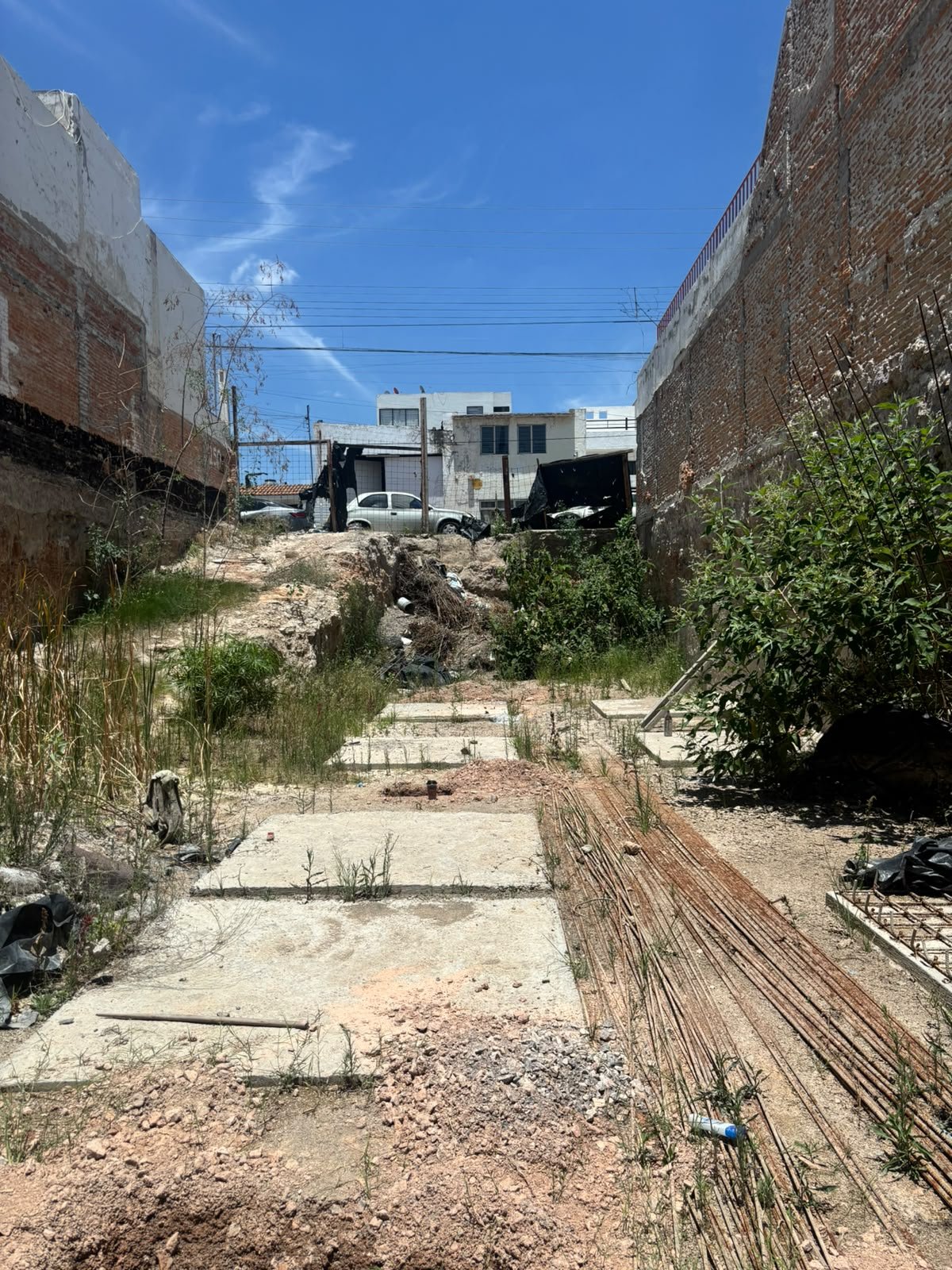 Un terreno en construcción con suelo de tierra y piedras, con algunos hierbajos creciendo y un par de rebaras de acero, rodeado por muros de ladrillo y paredes de concreto, con viviendas en el fondo y una línea de cables eléctricos bajo un cielo azul con algunas nubes.