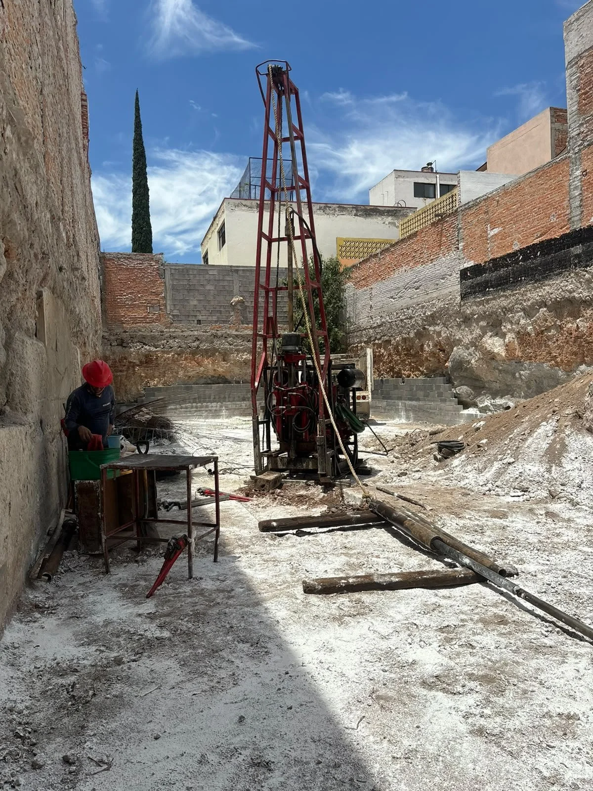 Construcción de una excavación en un entorno urbano con maquinaria pesada y un trabajador con casco rojo.