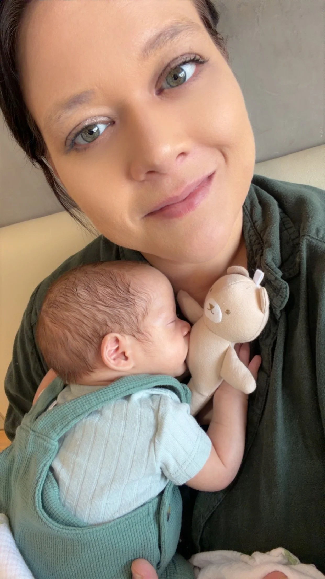 A woman with short dark hair and gray eyes holding a sleeping baby who is hugging a stuffed animal, in a close-up indoor setting.