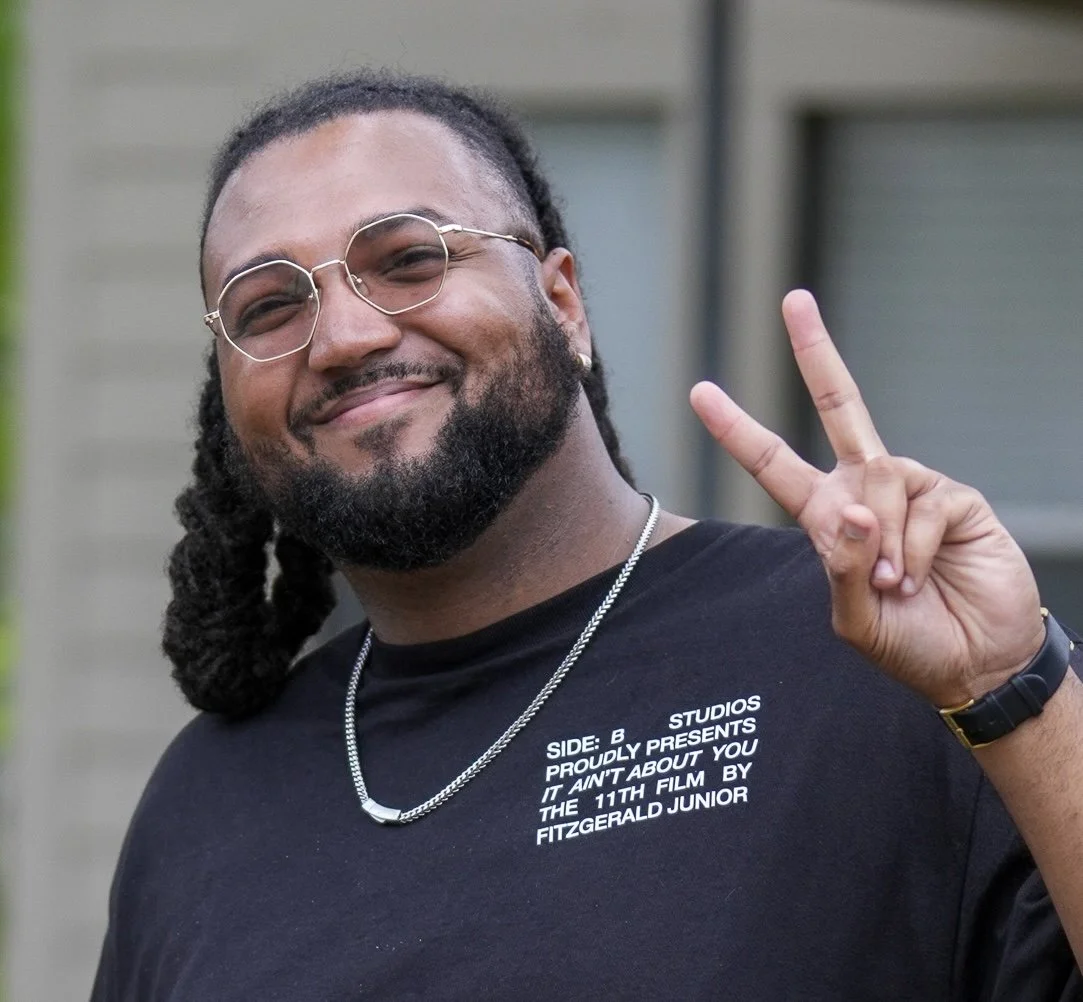 A man with glasses, a beard, and dreadlocks, smiling and making a peace sign with his hand.