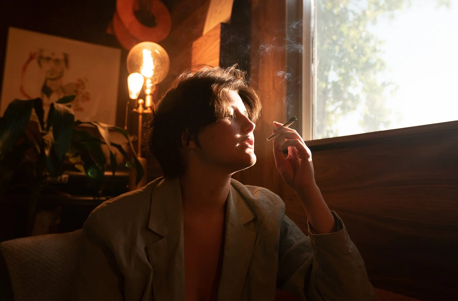 A woman sitting by a window, smoking a cigarette, with sunlight illuminating her face from outside, in a warmly lit room with artwork and a lamp in the background.