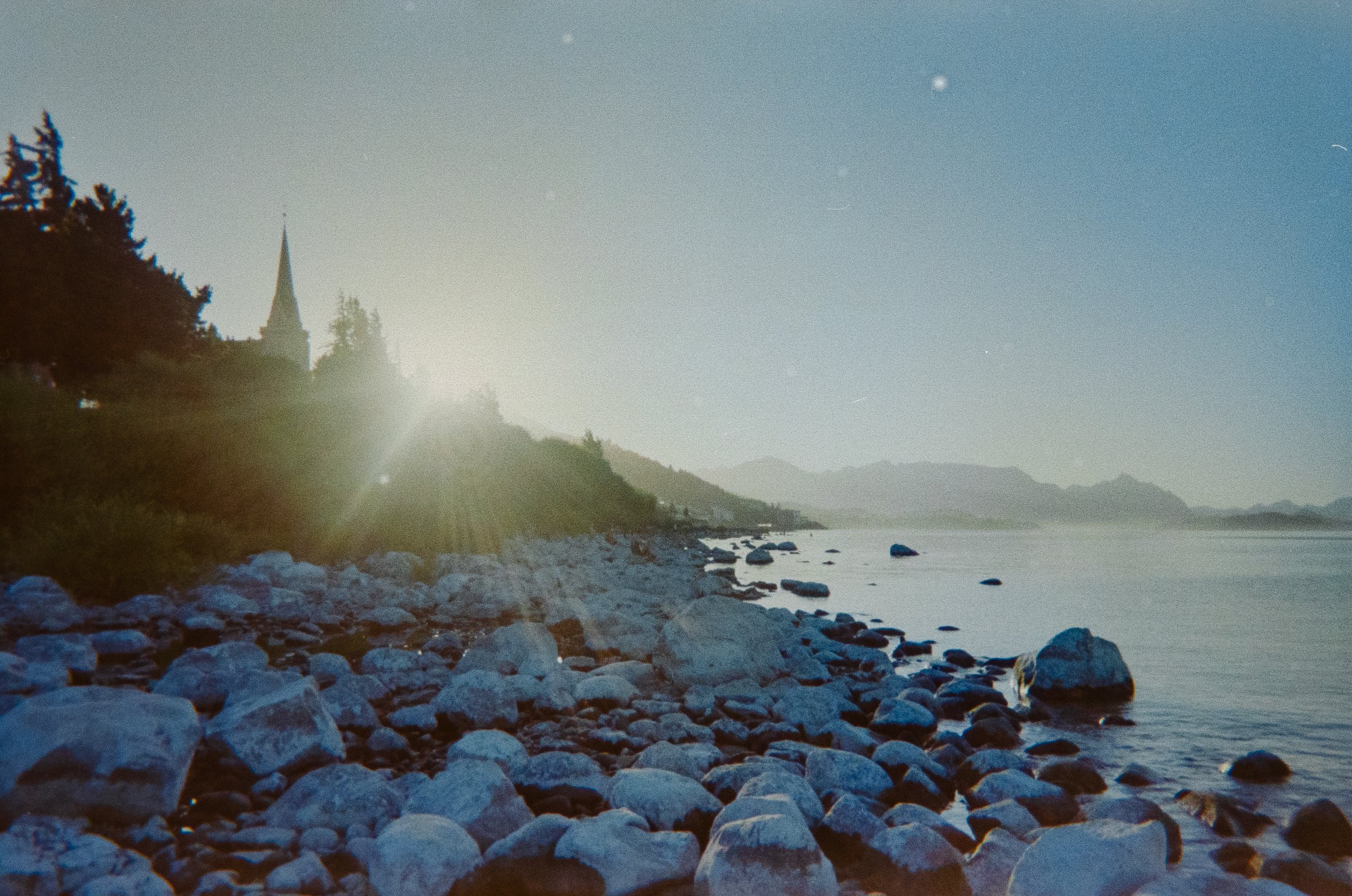 Scenic view of a river with a rocky shoreline, trees on a hill, and a church steeple in the background with sun shining behind it, creating a bright glare in the image.