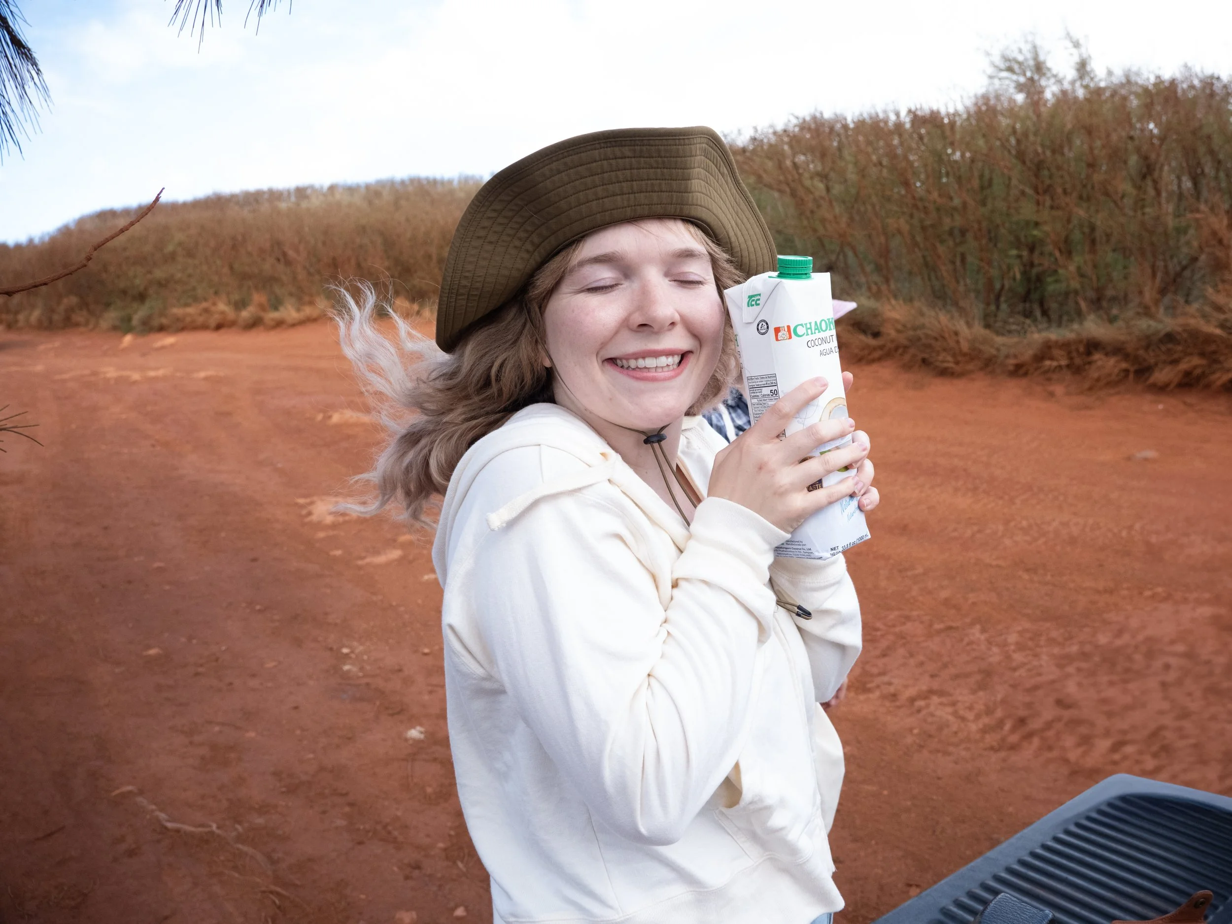 A young woman with curly blonde hair wearing a wide-brimmed hat and white jacket holding a carton of coconut water, smiling with closed eyes outdoors on a reddish-orange dirt path with trees in the background.