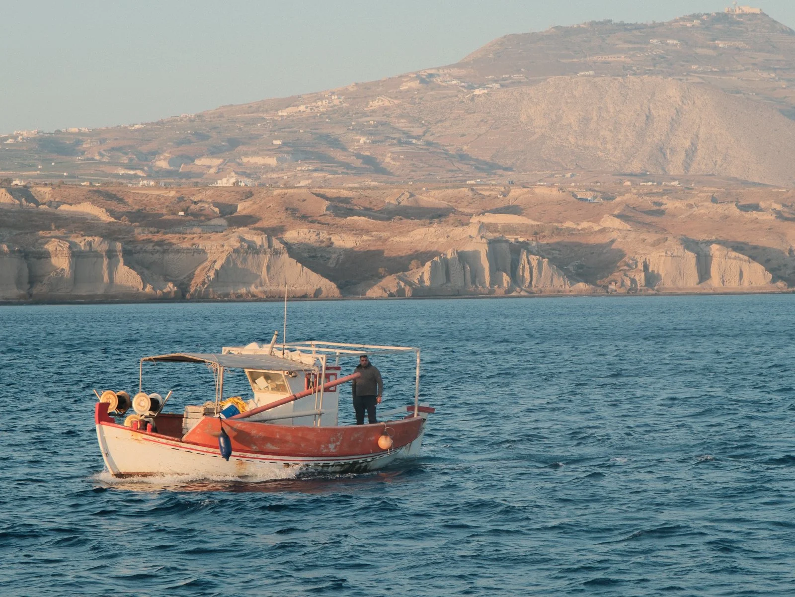 A small boat on the water with a man standing on it, with a hilly landscape in the background.